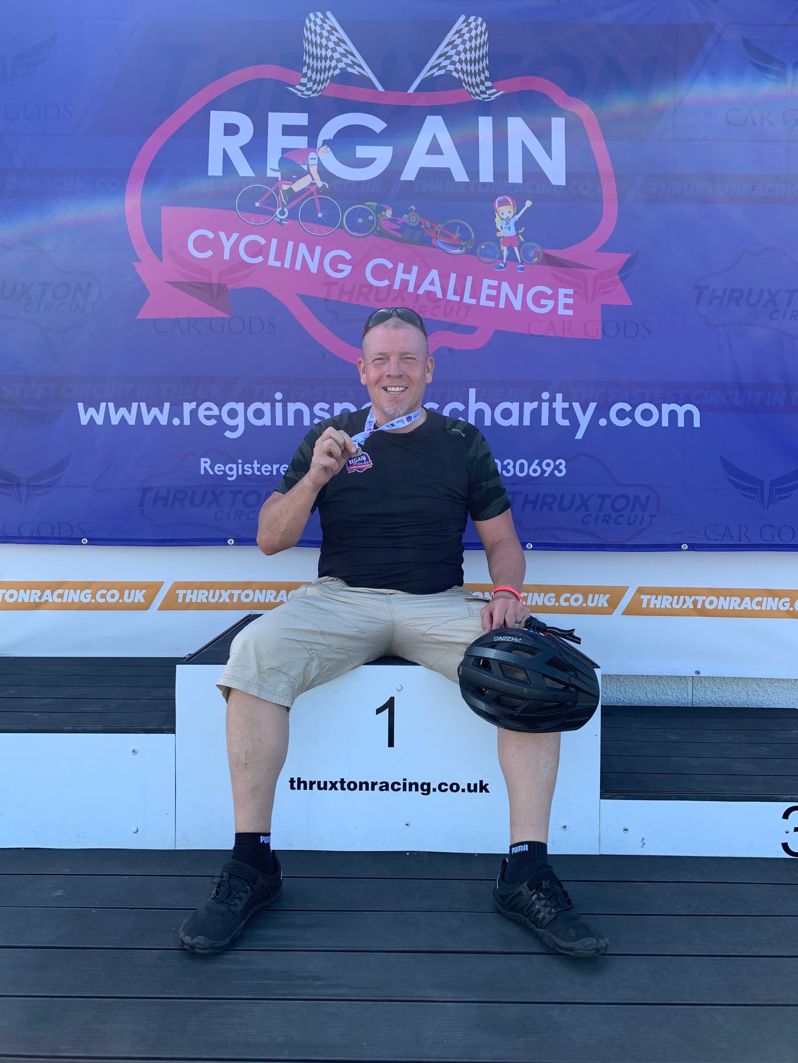 A man sits on a podium holding a medal and a bicycle helmet in front of a "Regain Cycling Challenge" banner. He is wearing a black shirt and shorts, and is smiling at the camera.