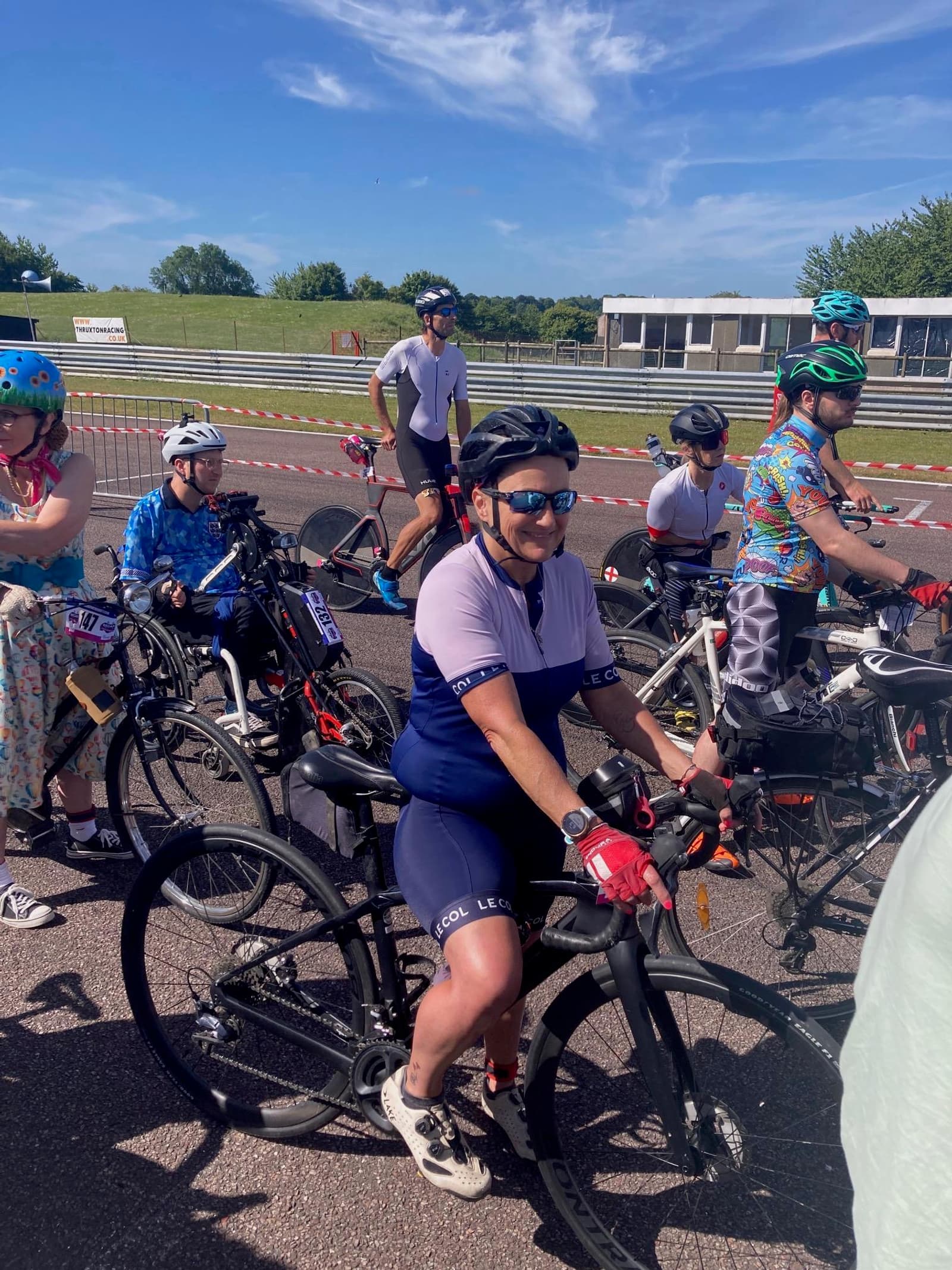 Cyclists gathered on a sunny day, preparing for a ride on a racetrack. They wear helmets and various cycling attire. The background features a grassy area and a building.