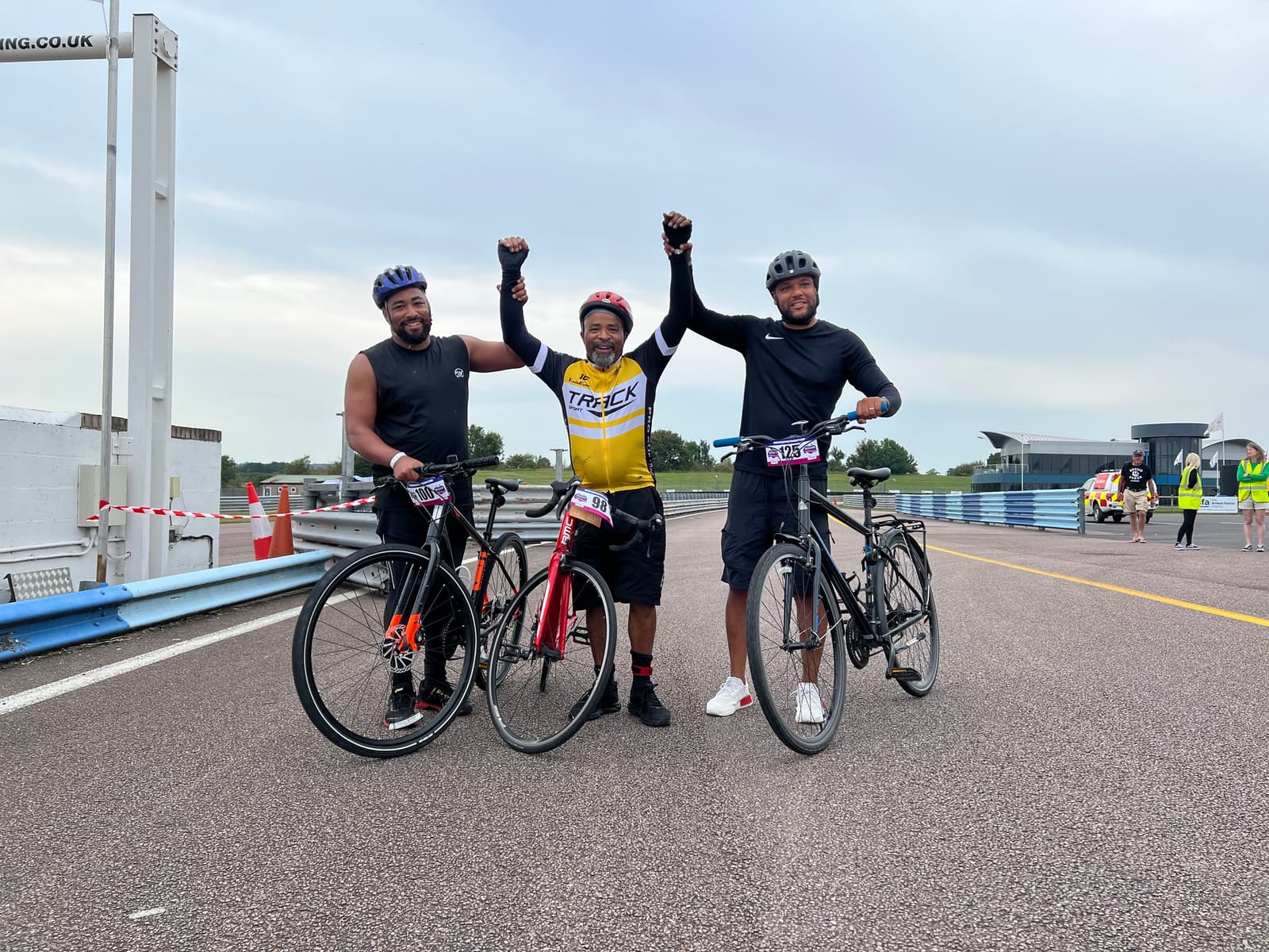 Three cyclists stand on a track, holding hands triumphantly. The person in the center wears a yellow jersey, while the others wear black. Each is standing with their bike. The background shows a cloudy sky and race track facilities.