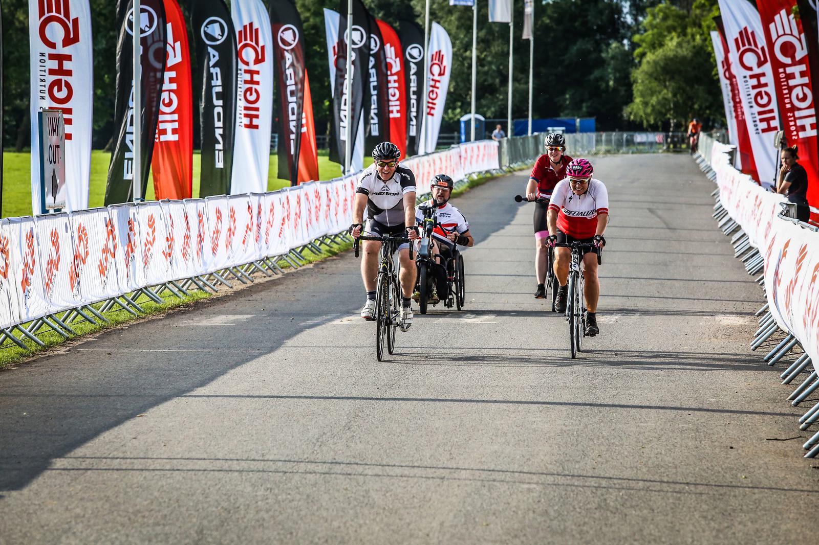 A group of athletes participating in a cycling event ride down a road lined with sponsor banners. The road is bordered by barriers, and greenery is visible in the background. One athlete in a handcycle is flanked by cyclists on traditional bicycles.