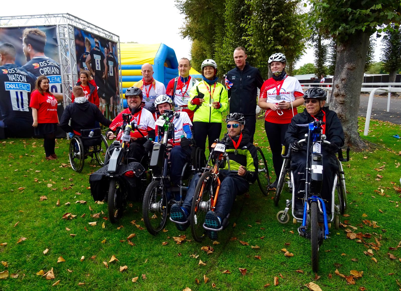 A group of people, some in wheelchairs, pose together on grass by a tree. They are wearing various gear and accessories, including cycling helmets and red uniforms. A large poster and an inflatable structure are visible in the background.