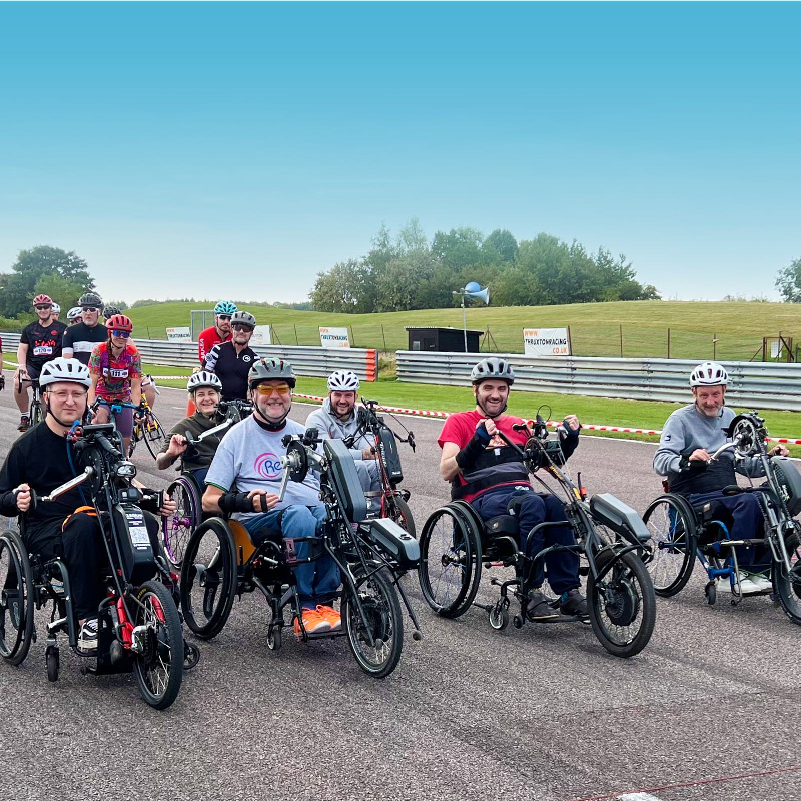A group of eight people with various disabilities are smiling and riding adaptive bicycles on a racetrack. They are wearing helmets, and the background features a clear, blue sky, green grass, a few trees, and a safety barrier along the track's edge.