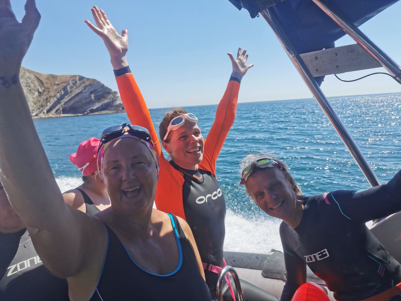 Three smiling individuals in wetsuits are on a boat in sunny conditions with the ocean and part of a rocky coastline visible in the background. The person in the middle has their arms raised joyfully, while the others also display happy expressions.