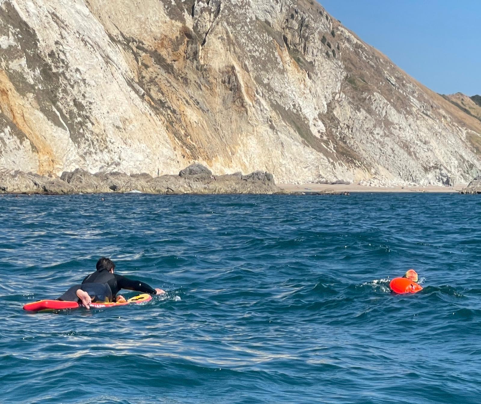 Two people wearing black wetsuits are in the ocean near a rocky shoreline. One person is lying on a red surfboard paddling, and the other is swimming nearby with a bright orange floatation device. The water is a deep blue, and the rocky cliff is partially sunlit.