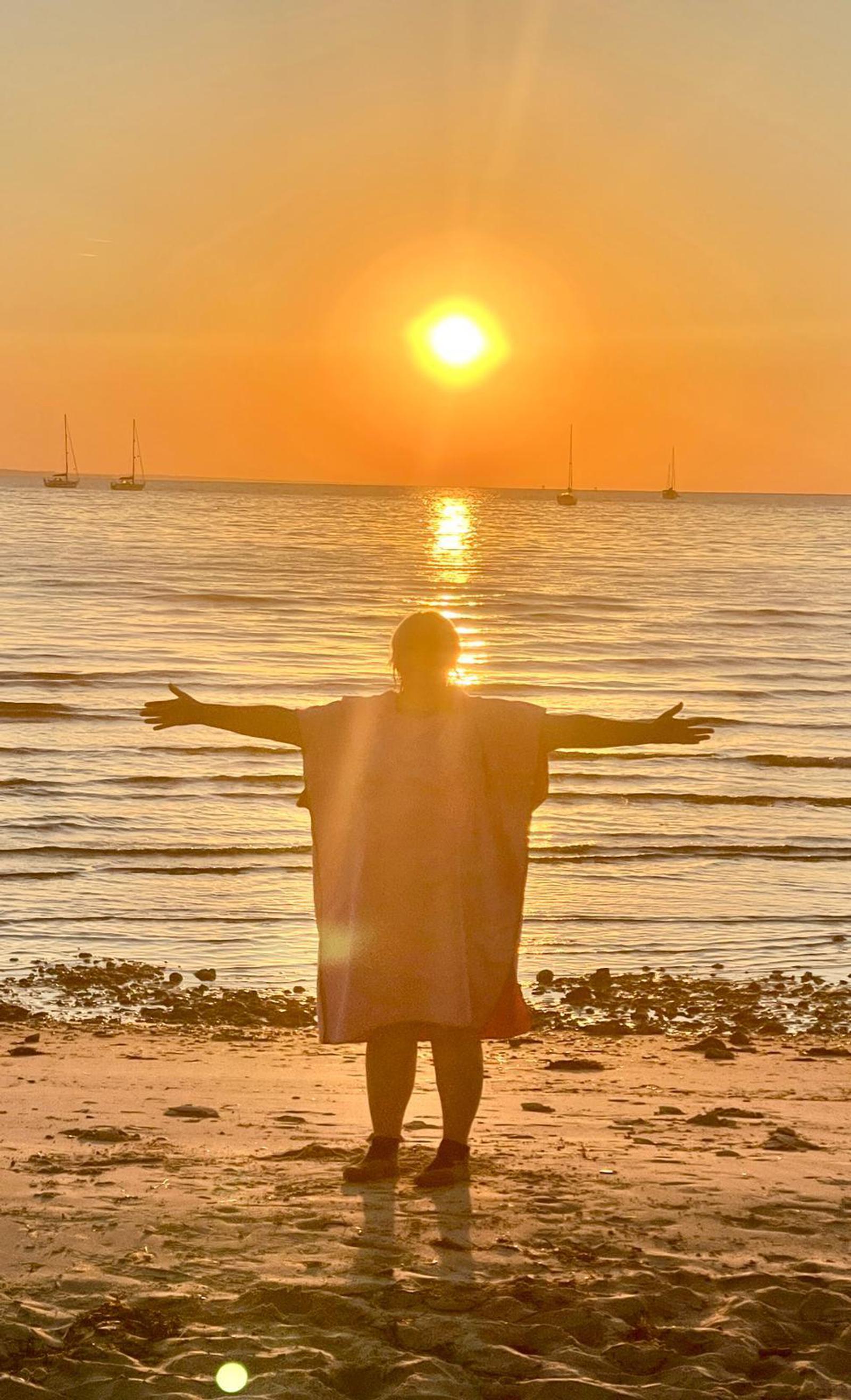 A person stands on a beach facing the ocean at sunset, arms outstretched. The sun is low in the sky, casting a golden glow over the scene. Several sailboats are visible on the horizon, and gentle waves lap at the pebbled shore.