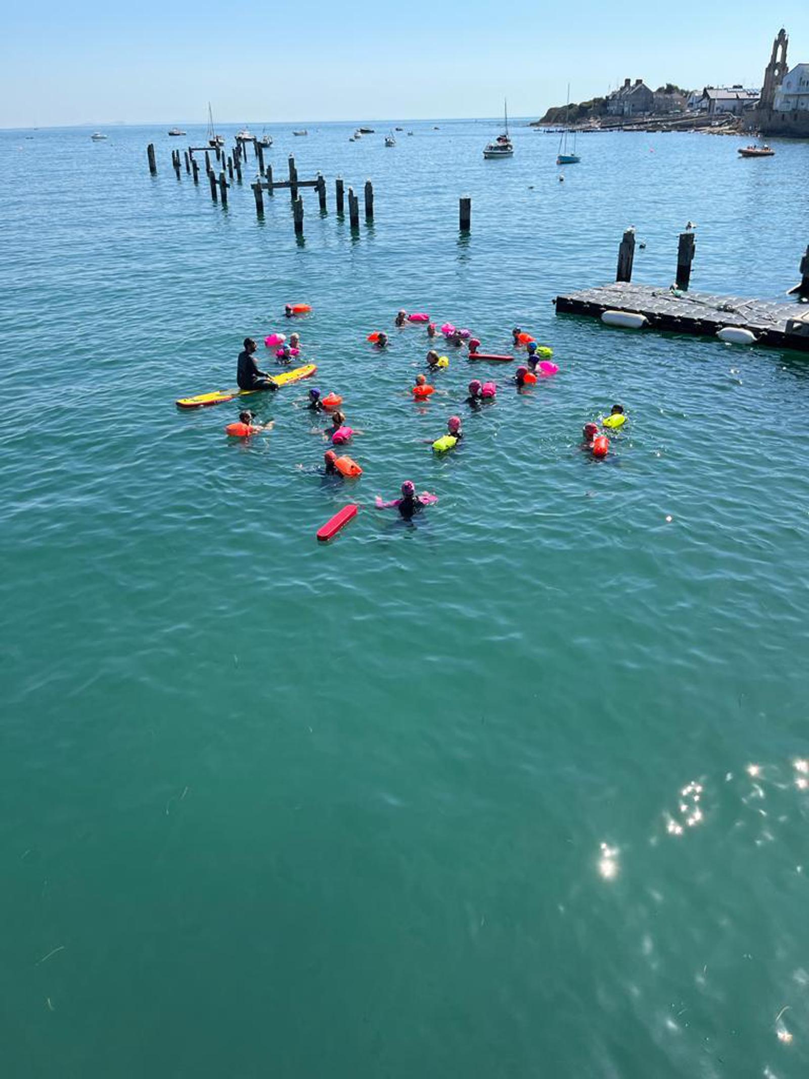 A group of swimmers with colorful floatation devices are spread out in a clear, calm sea near a weathered pier. A few small boats are anchored nearby, and a rocky shoreline with buildings is visible in the background under a bright, clear sky.