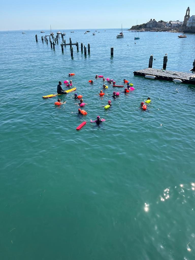 A group of swimmers with colorful floatation devices are spread out in a clear, calm sea near a weathered pier. A few small boats are anchored nearby, and a rocky shoreline with buildings is visible in the background under a bright, clear sky.