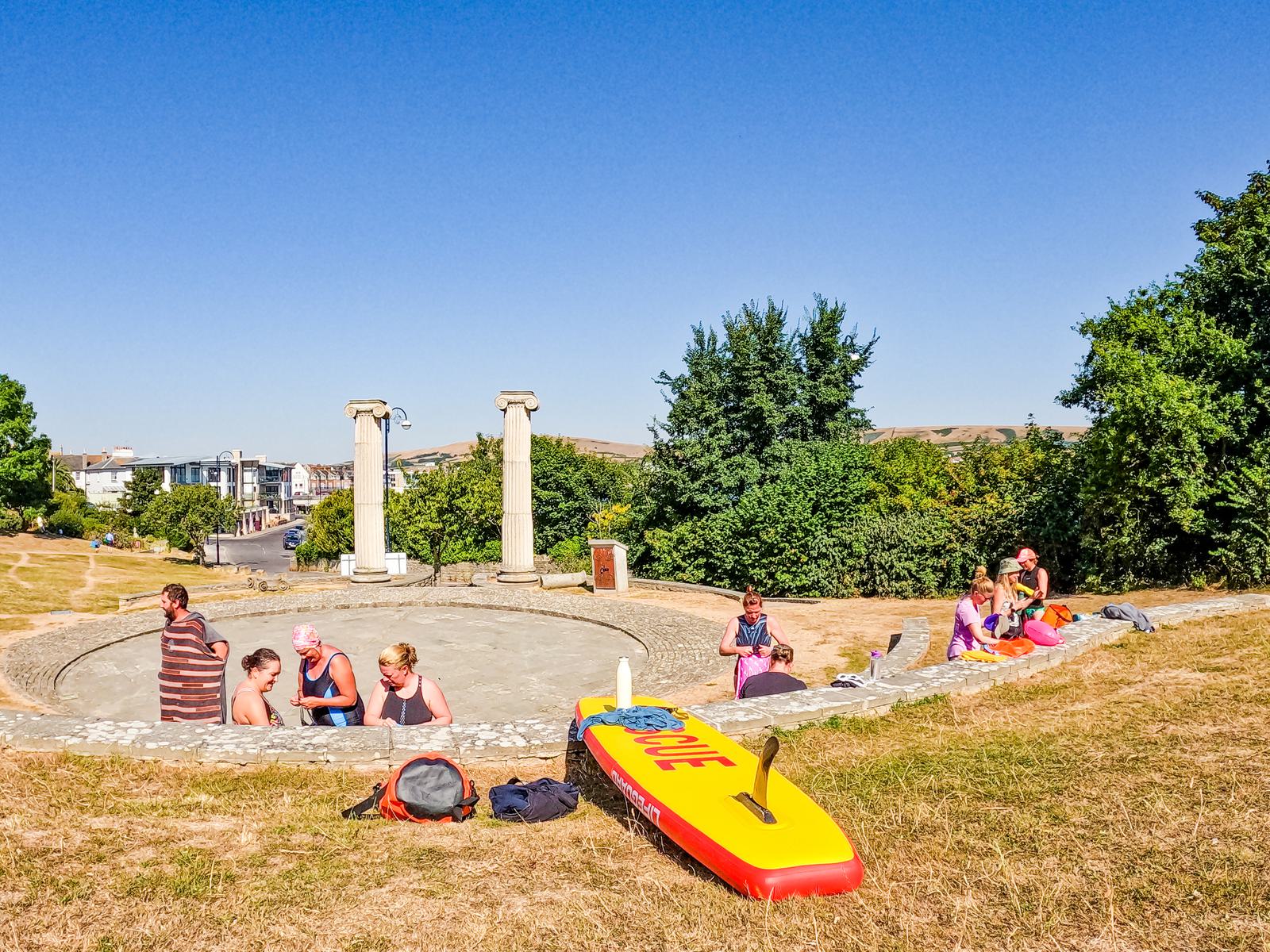 A group of people sitting and relaxing in a park on a sunny day. There's a yellow and red rescue board in the foreground, and two stone pillars in the background. Some trees and bushes are surrounding the area, with a few buildings visible in the distance.
