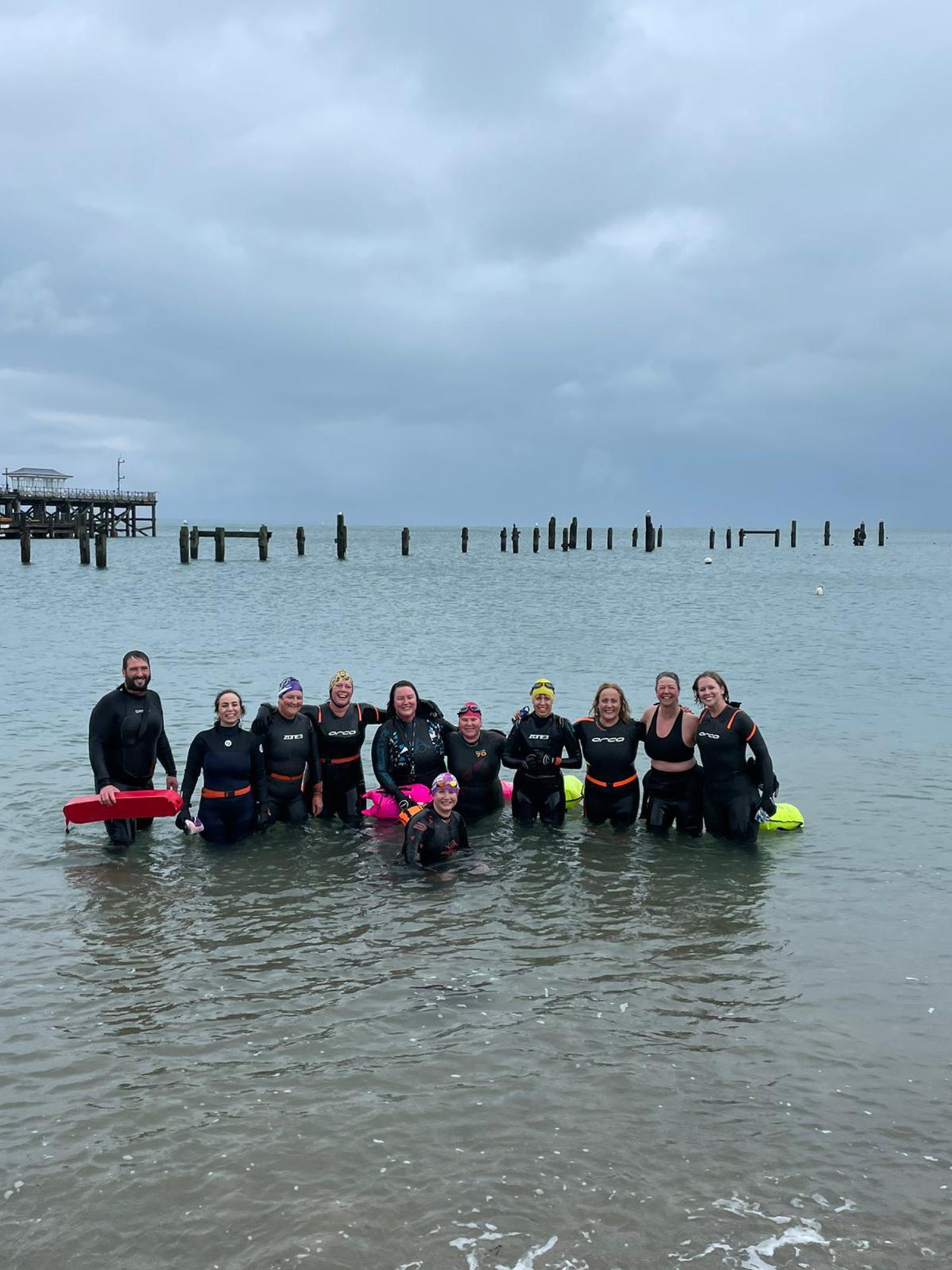 A group of people wearing wetsuits stand together in shallow ocean water for a photograph. The sky is cloudy, and remnants of an old pier can be seen in the background. Some group members hold flotation devices.