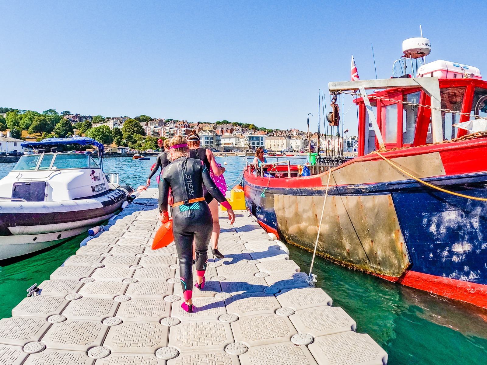 A group of people in wetsuits carrying orange flotation devices walks along a floating dock next to colorful boats. The dock is surrounded by clear blue water, and a coastal town with houses and greenery is visible in the background under a clear sky.