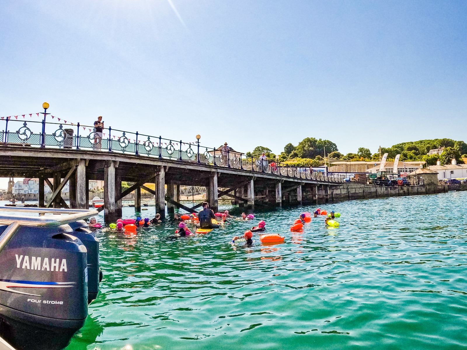 A group of swimmers in colorful caps and buoyancy aids float in a blue body of water under a sunny sky, near a wooden pier with people standing on it. A Yamaha boat engine is visible in the foreground, and there are buildings and trees in the background.