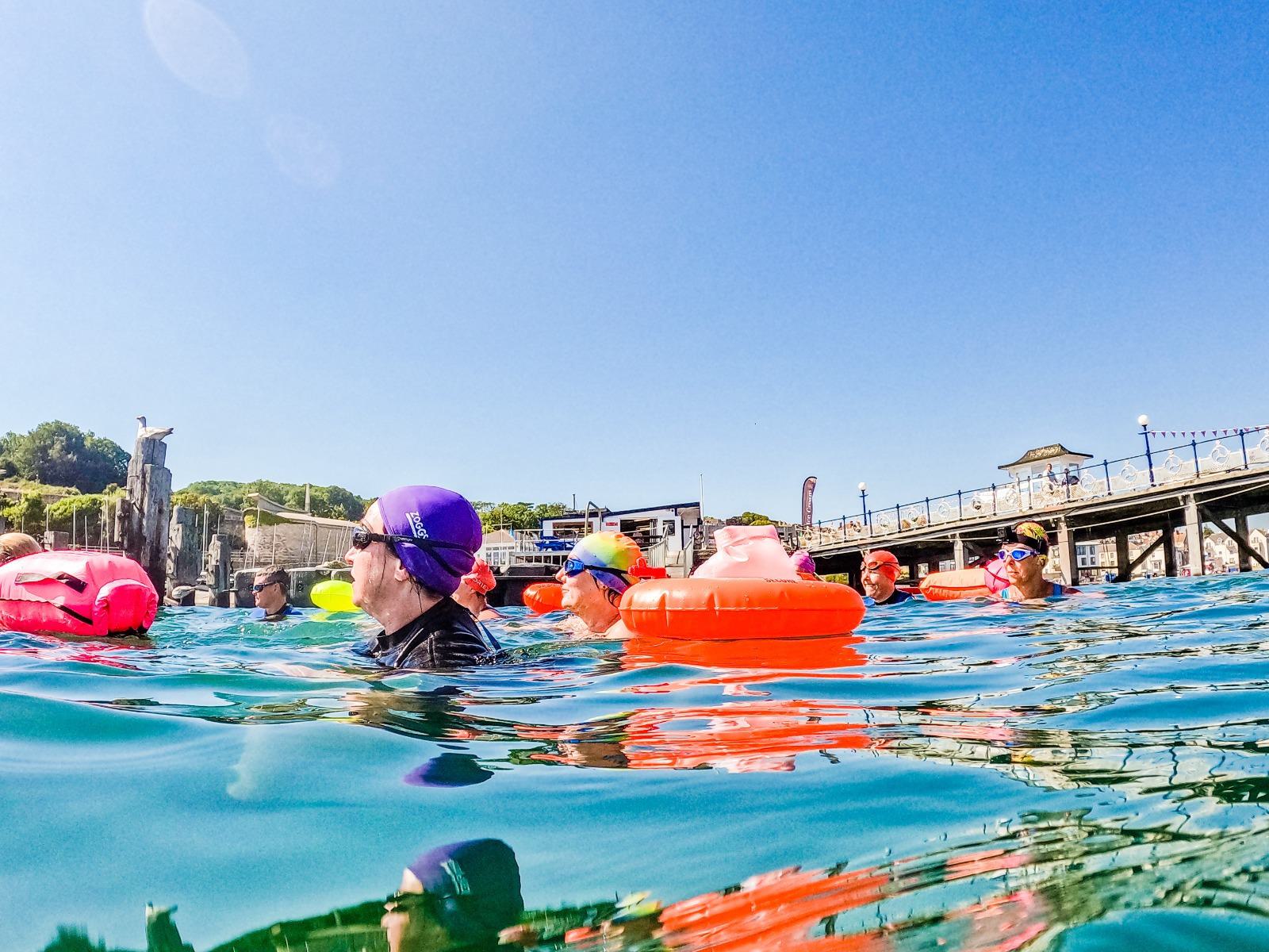 A group of swimmers with colorful swim caps and floats are in the water near a pier on a sunny day. The background shows a clear blue sky and a structure on the pier. Some people are partially submerged, while others float near the surface.