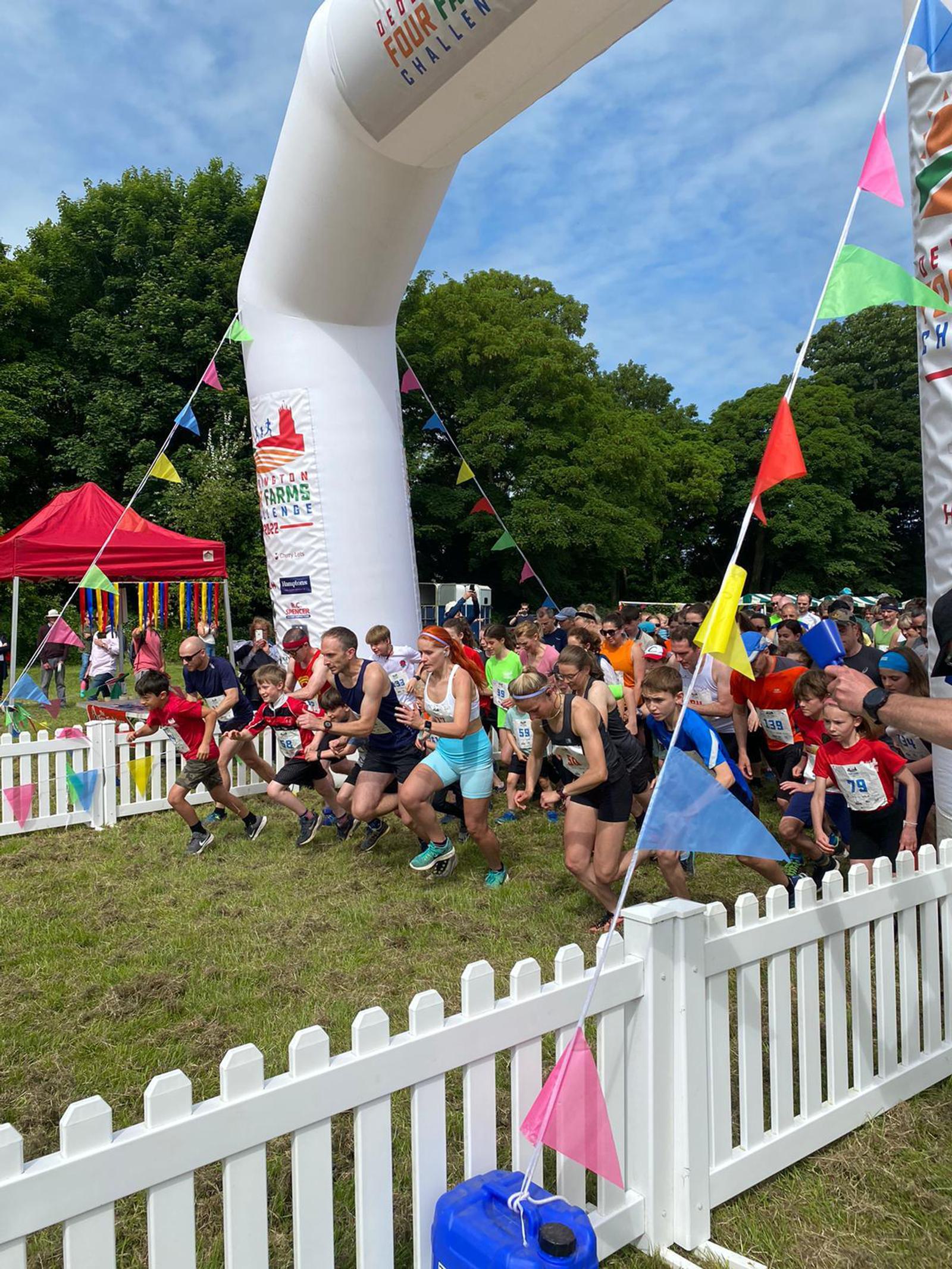Runners of various ages and genders start a race at an outdoor event. They run past a start line marked by an inflatable arch adorned with bunting and a banner. Spectators stand behind a white fence, and tents are visible in the background amidst trees.