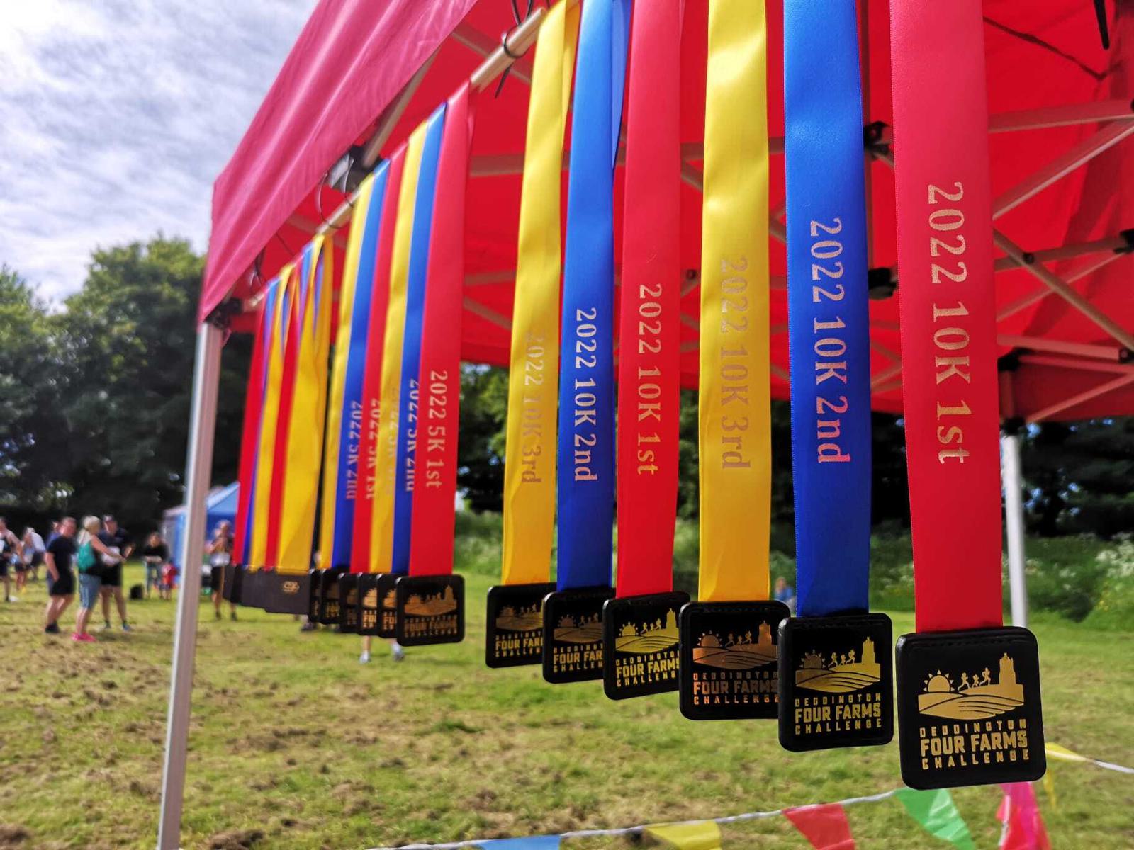 Brightly colored ribbons, each with a medal, hang from a red canopy at an outdoor event. The ribbons display various inscriptions like "2022 10K 1st," "2022 10K 2nd," and "2022 5K 1st," and the medals read "Four Farms Challenge." People and trees are visible in the background.
