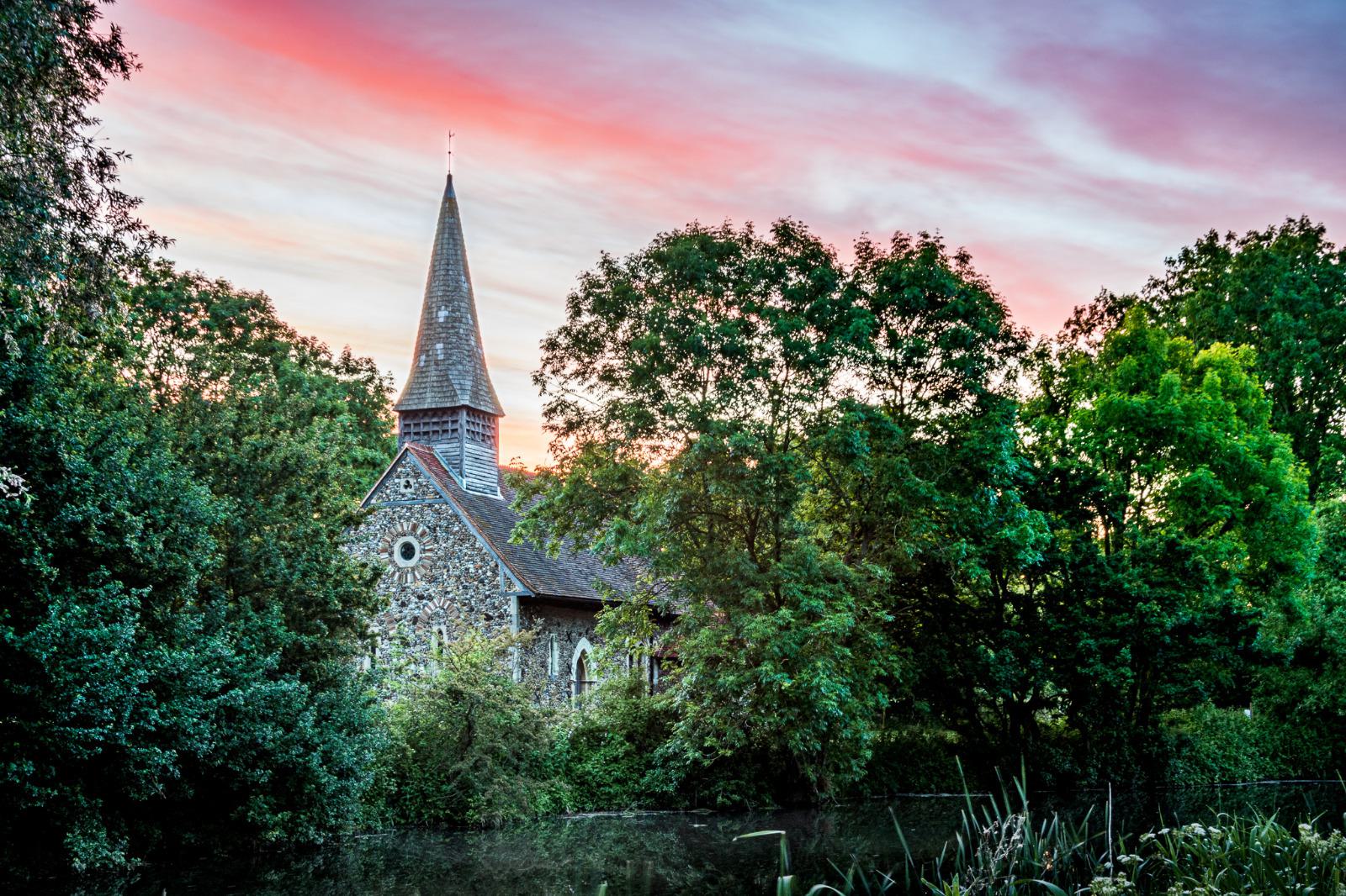 A stone church with a tall, pointed steeple is partially obscured by lush green trees. The sun is setting, casting a colorful pink and orange hue across the sky, reflecting beautifully in a pond in the foreground.