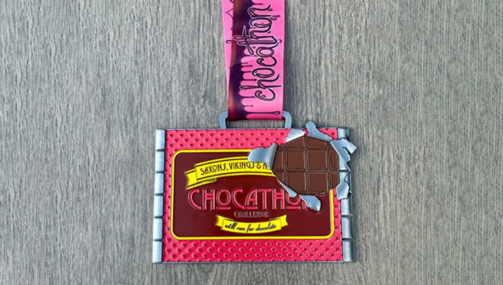 A pink and silver "Chocathon" race medal with a chocolate bar design and a bitten corner, attached to a pink ribbon on a gray wooden surface.