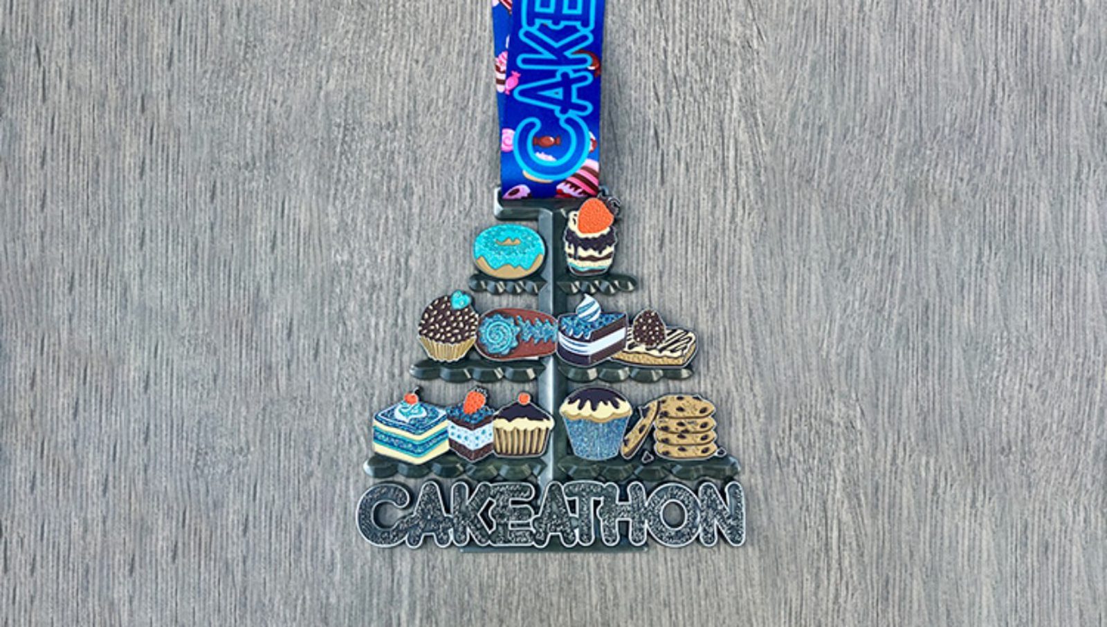 A medal with the word "CAKEATHON" at the bottom, decorated with images of cakes, cupcakes, and cookies, hangs from a blue ribbon with "CAKE" written on it, placed on a gray wooden surface.
