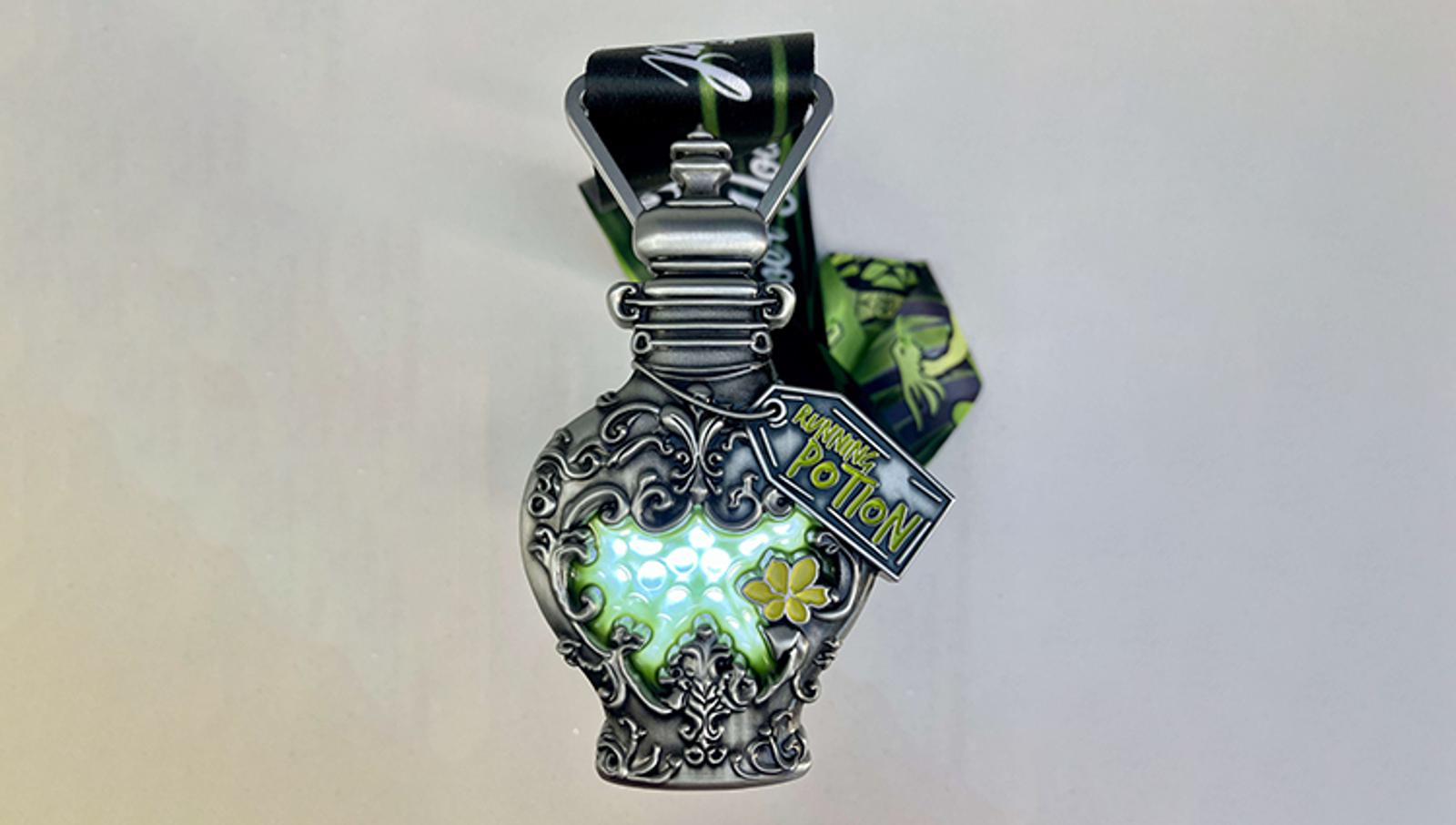 A decorative, ornate medal shaped like a potion bottle with a glow effect, attached to a black and green ribbon. The medal features intricate silver designs and a small tag that reads "5K Finisher Potion.