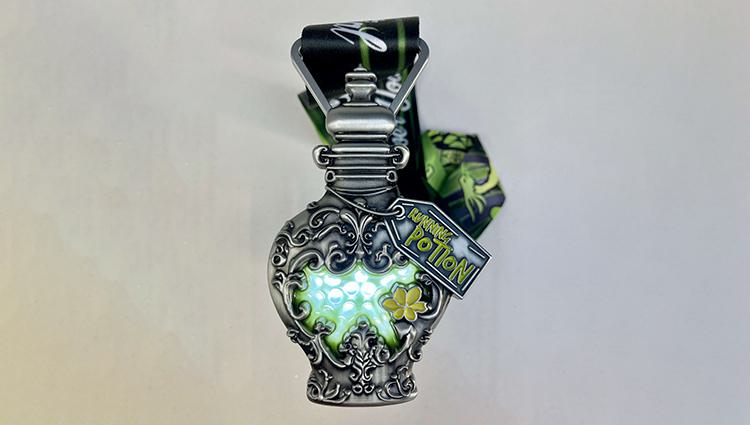 A decorative, ornate medal shaped like a potion bottle with a glow effect, attached to a black and green ribbon. The medal features intricate silver designs and a small tag that reads "5K Finisher Potion.