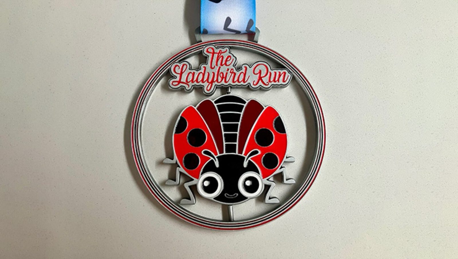 A medal with a cartoon ladybird in the center and the text "The Ladybird Run" above it, attached to a blue and white ribbon, displayed on a plain light surface.