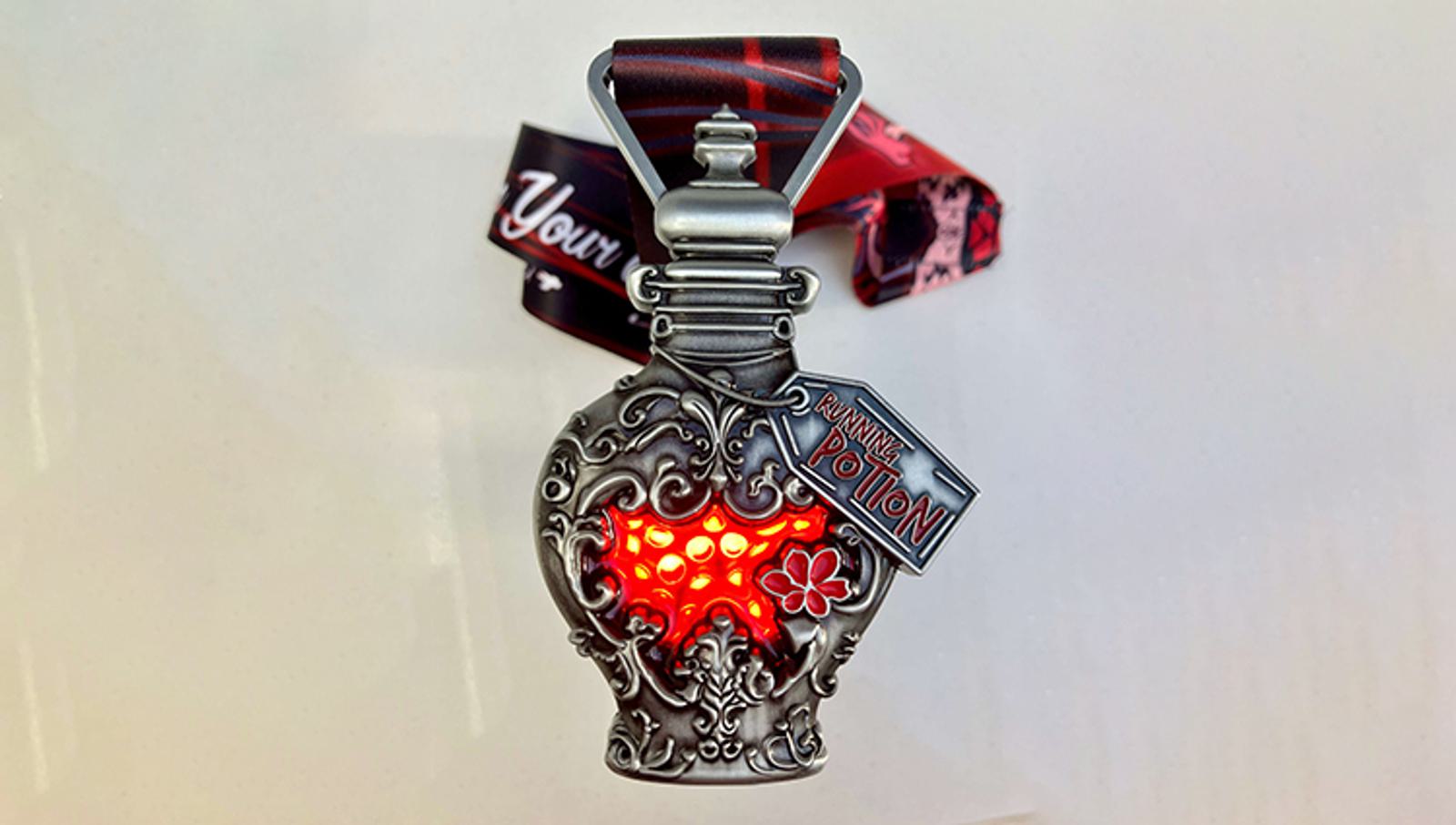 A decorative medal in the shape of an ornate potion bottle with a glowing red center. It has intricate floral designs and a tag reading "RUN MAGICAL POTION." The attached ribbon has a mix of red and black colors.