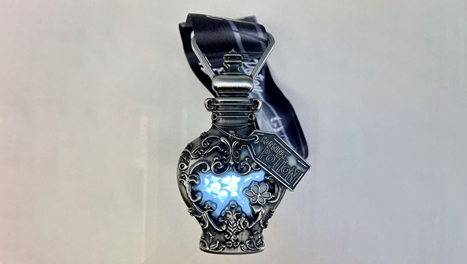 A silver decorative medal shaped like a potion bottle with intricate designs and a tag reading "10K" attached. The bottle has a glowing blue light inside. A black and silver ribbon is attached to the top.