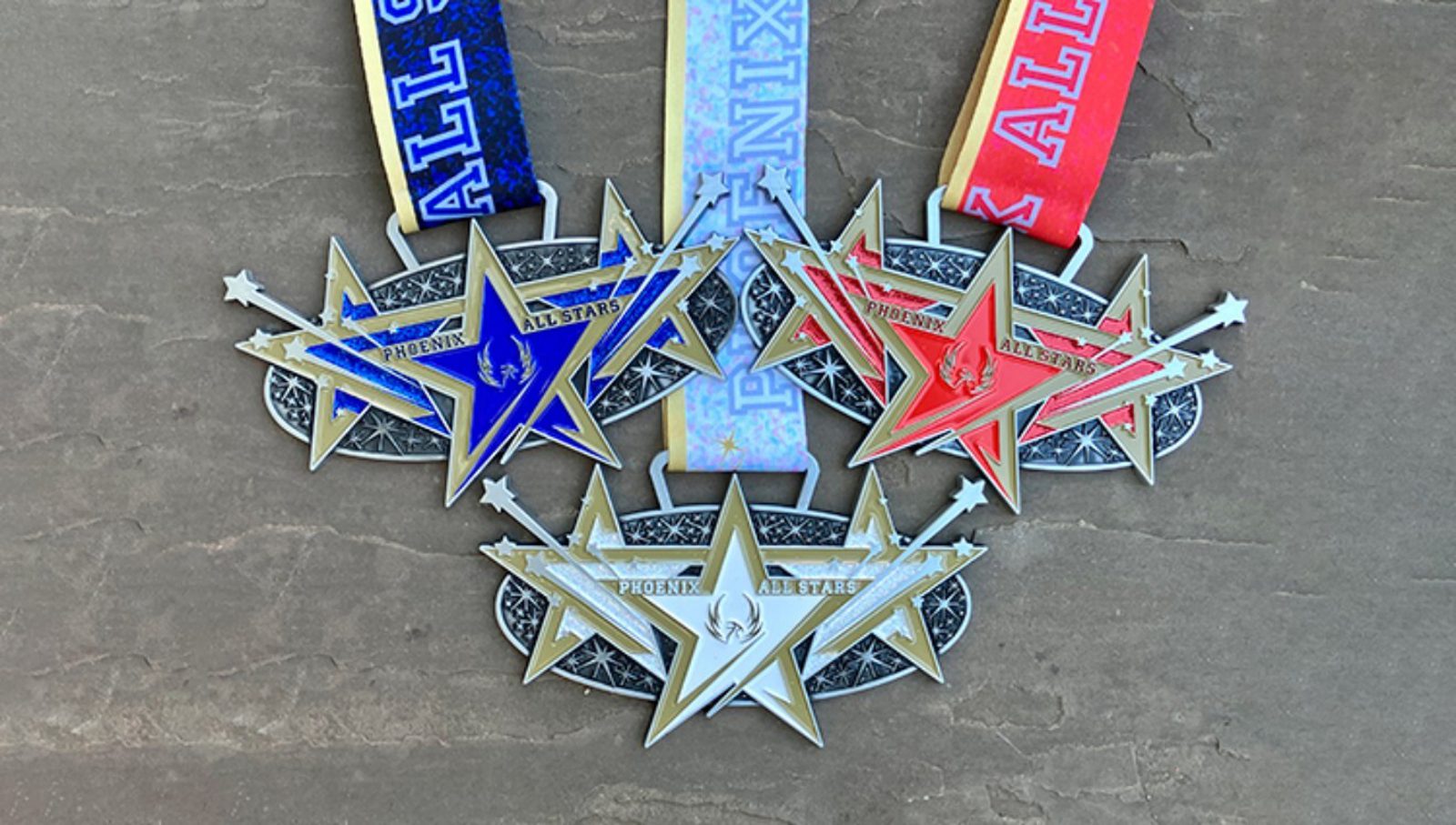 Three star-shaped medals with "Phoenix Allstars" text are displayed side by side. The medals have blue, white, and red ribbons, and are set on a textured brown surface.