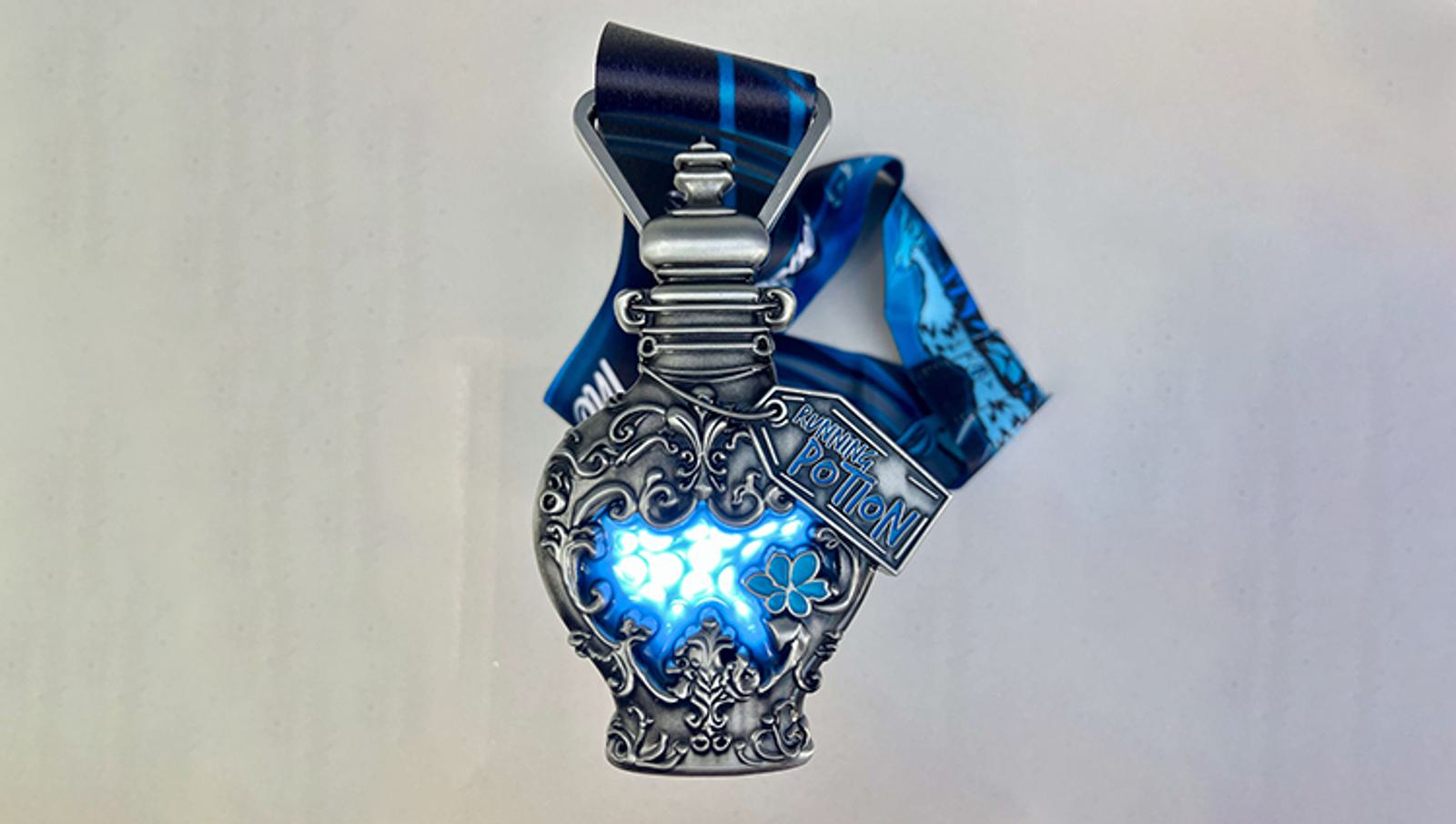 A decorative silver medal shaped like a potion bottle with intricate designs and a glowing blue center. It hangs from a blue ribbon. A small tag reads "Challenge Potion.