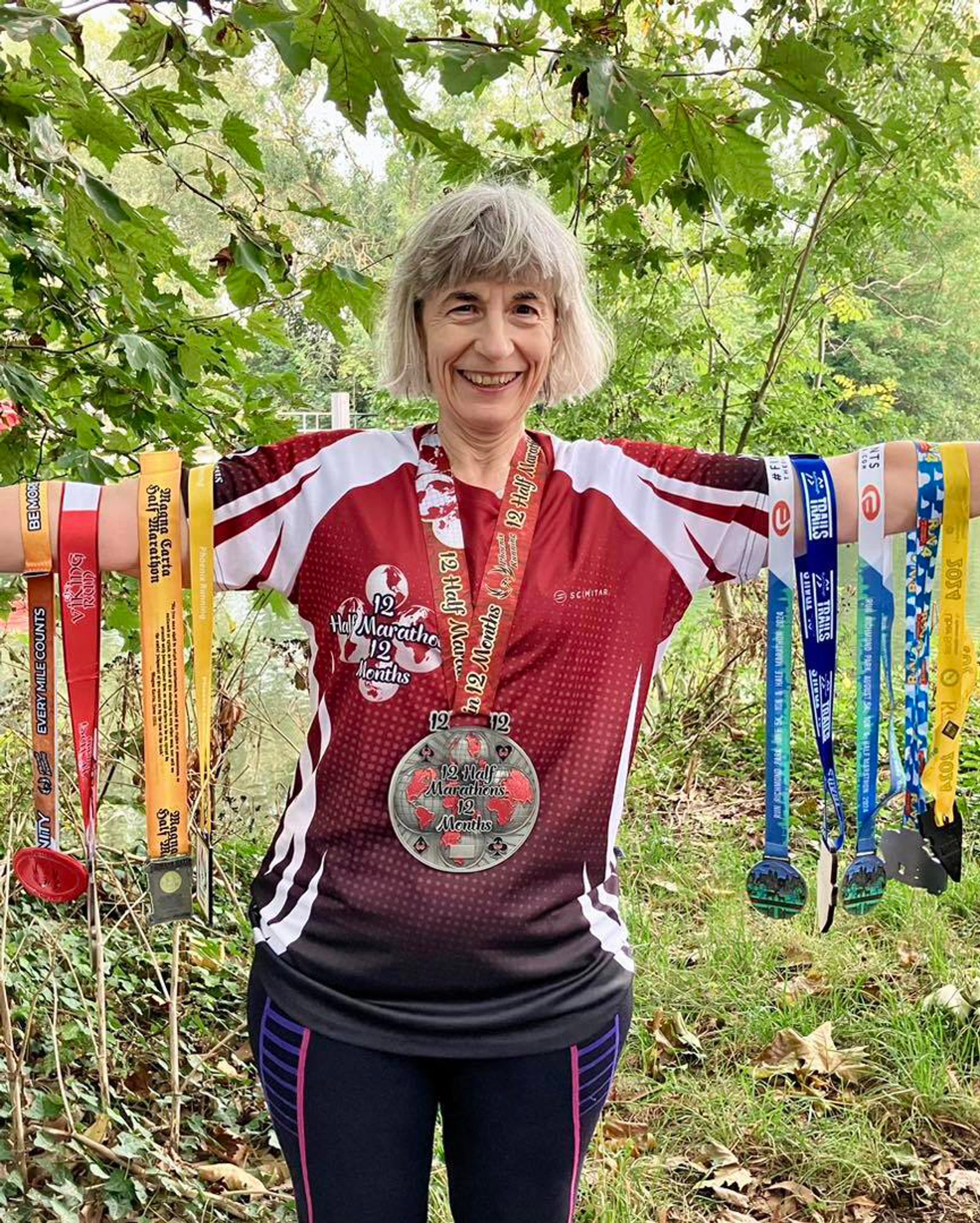 A person stands outdoors, smiling, while holding numerous colorful marathon medals on each arm. They wear a red and white sports shirt with a large medal hanging on their chest. Green trees and fallen leaves are visible in the background.