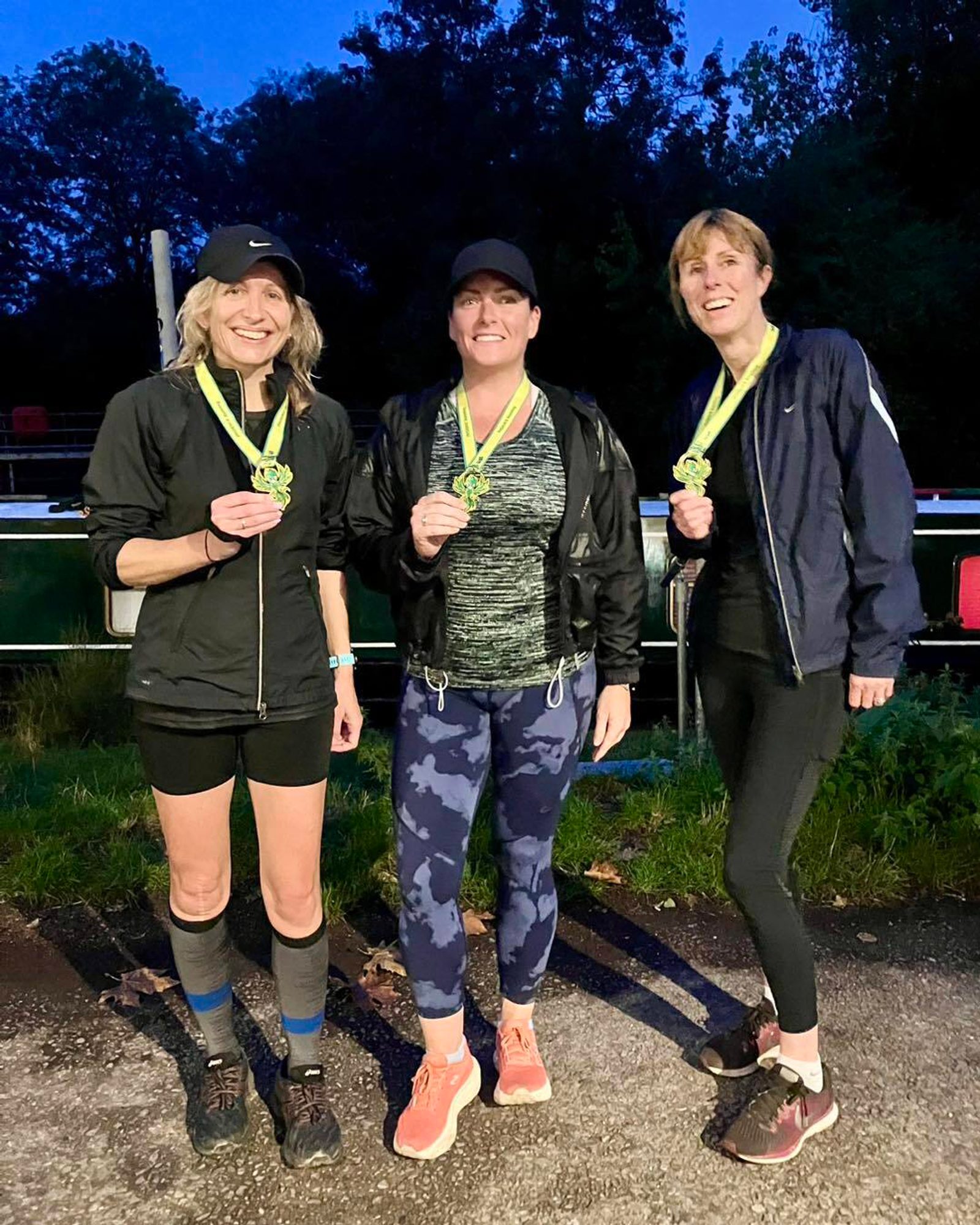 Three women wearing athletic clothing and caps stand outdoors, smiling and holding medals. The background features trees and a canal or river. It appears to be early morning or evening.