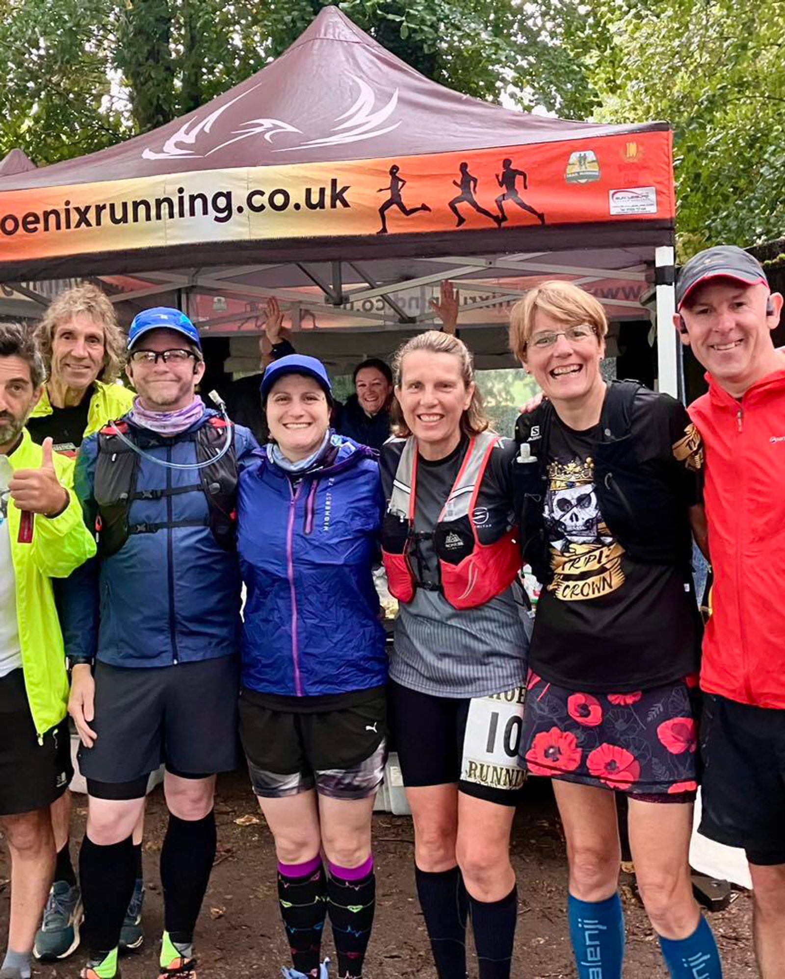 A group of runners poses together under a Phoenix Running tent. They are dressed in various athletic gear and appear happy and energetic after an event. Trees and a canopy are visible in the background.