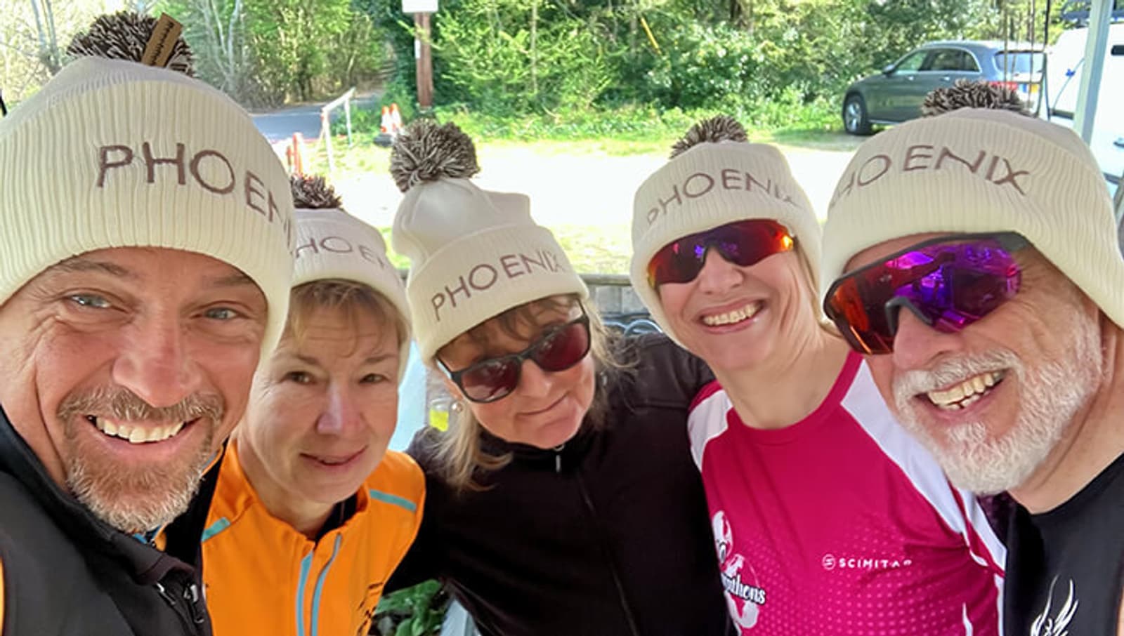 Five smiling adults wearing cream-colored "PHOENIX" beanies stand closely together outdoors, posing for a group selfie. Some are wearing sunglasses and colorful athletic clothing, with trees in the background.