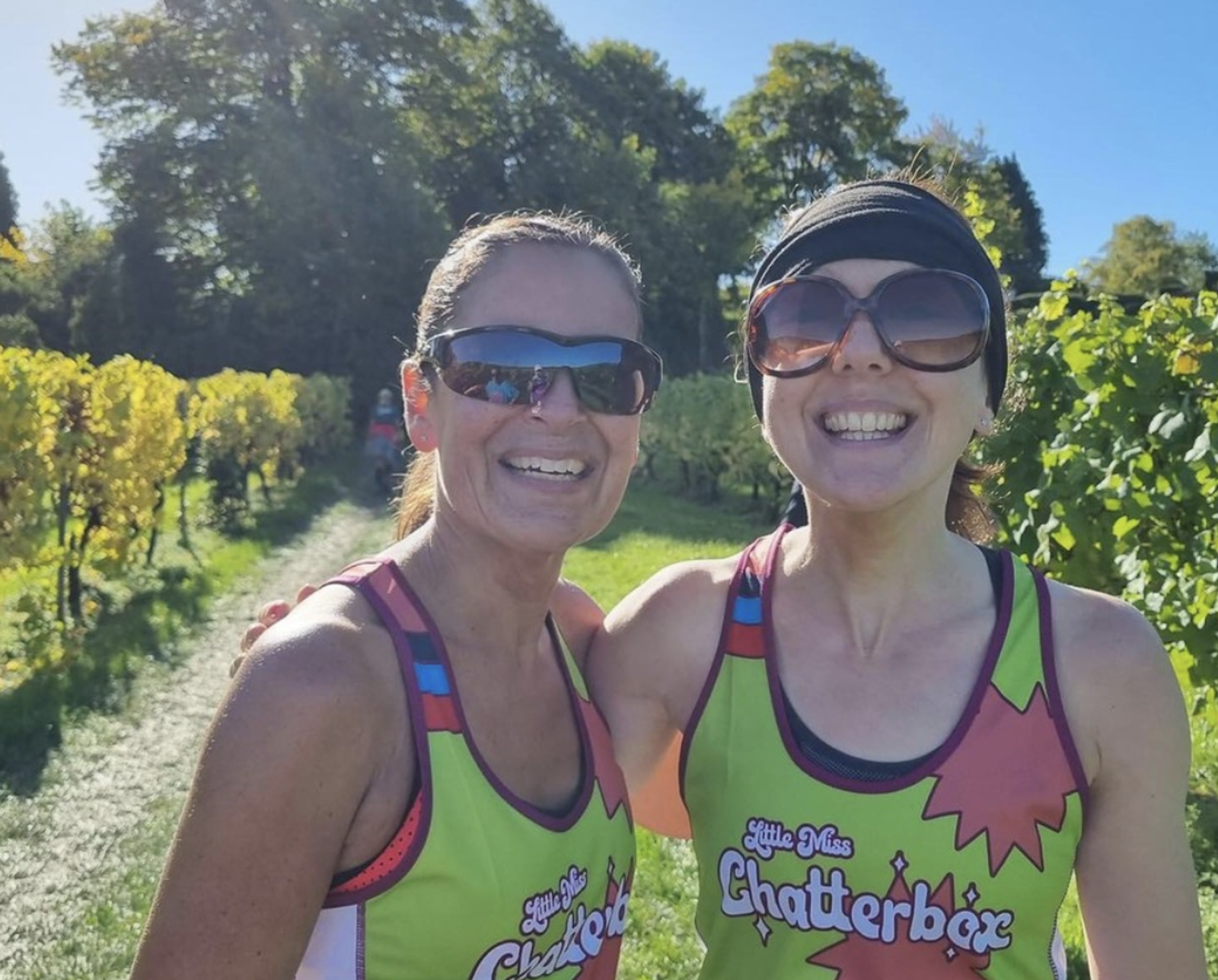 Two women wearing sunglasses and bright green tank tops with "Little Miss Chatterbox" text are smiling outdoors on a sunny day. They stand on a path between green vineyards with blue skies and trees in the background.