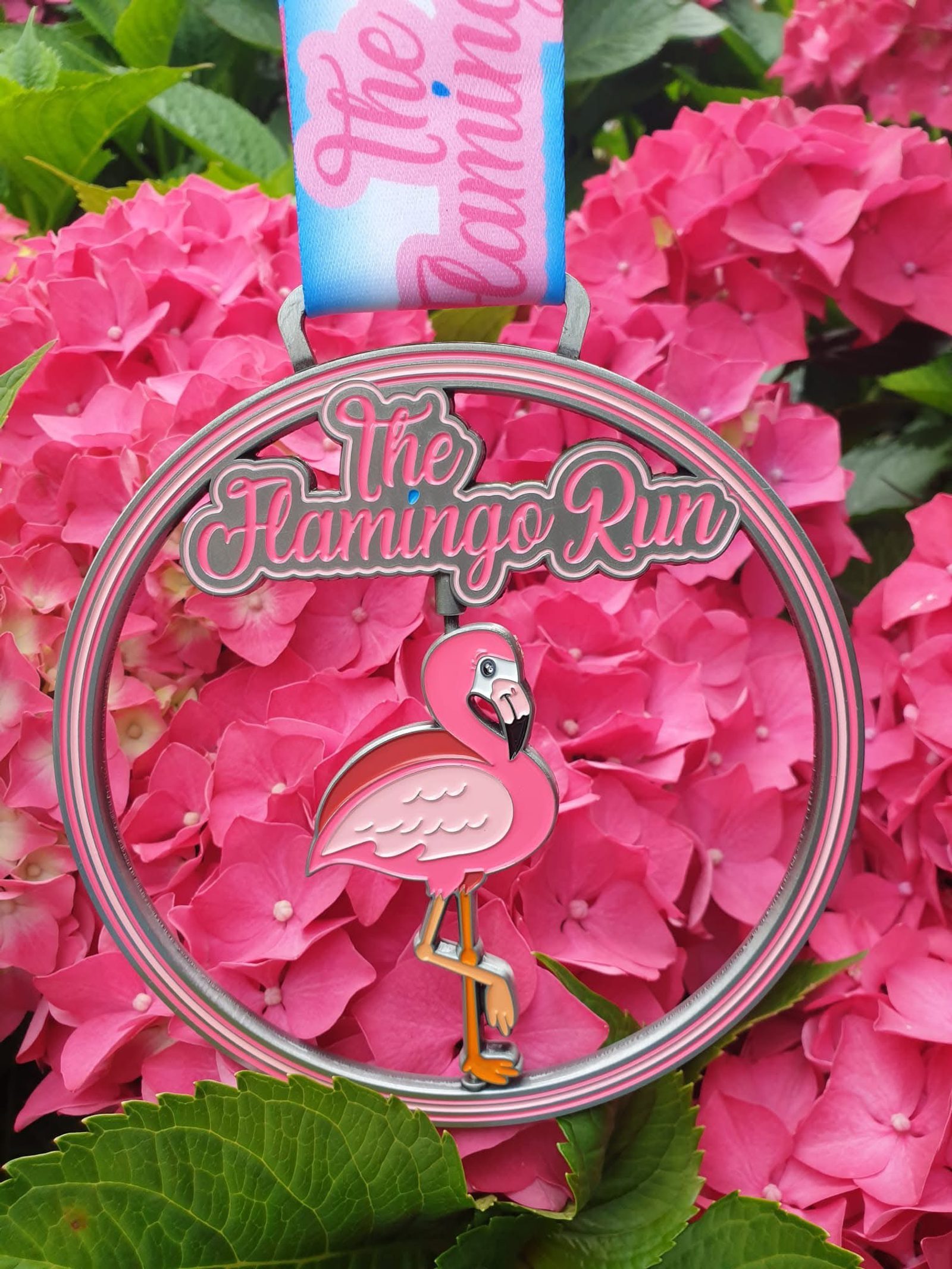 A medal with "The Flamingo Run" and a pink flamingo hangs in front of vibrant pink hydrangea flowers. The medal's ribbon is blue and pink with cursive writing.