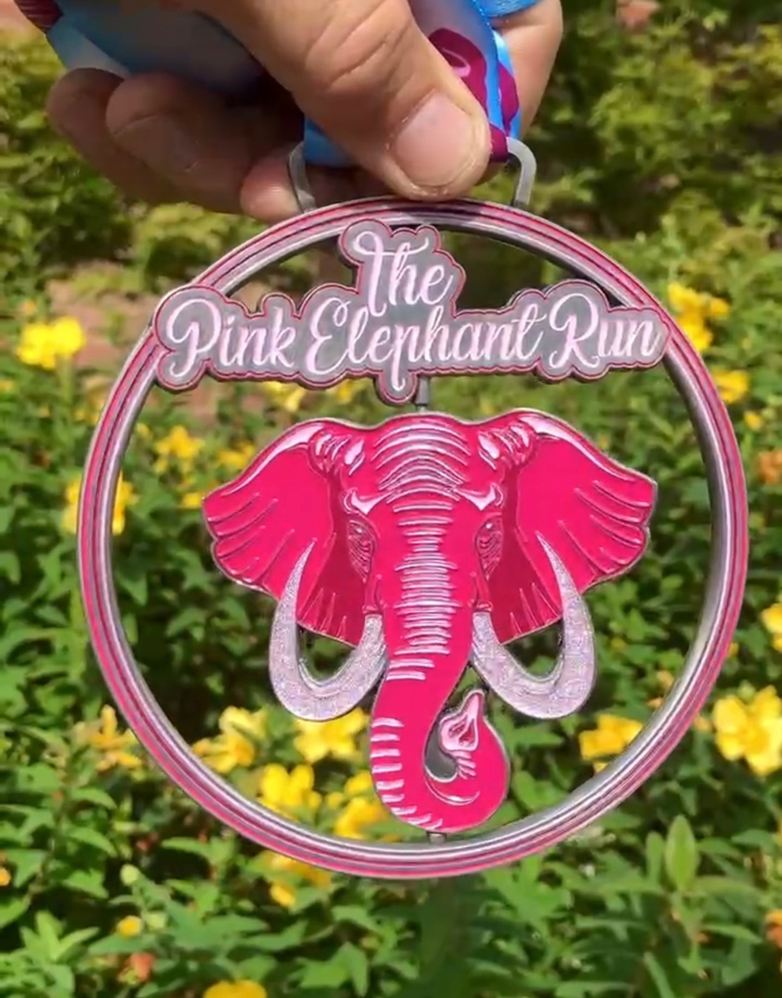 A hand holds a round medal for "The Pink Elephant Run," featuring a large pink elephant head in the center, with yellow flowers and green foliage in the blurred background.