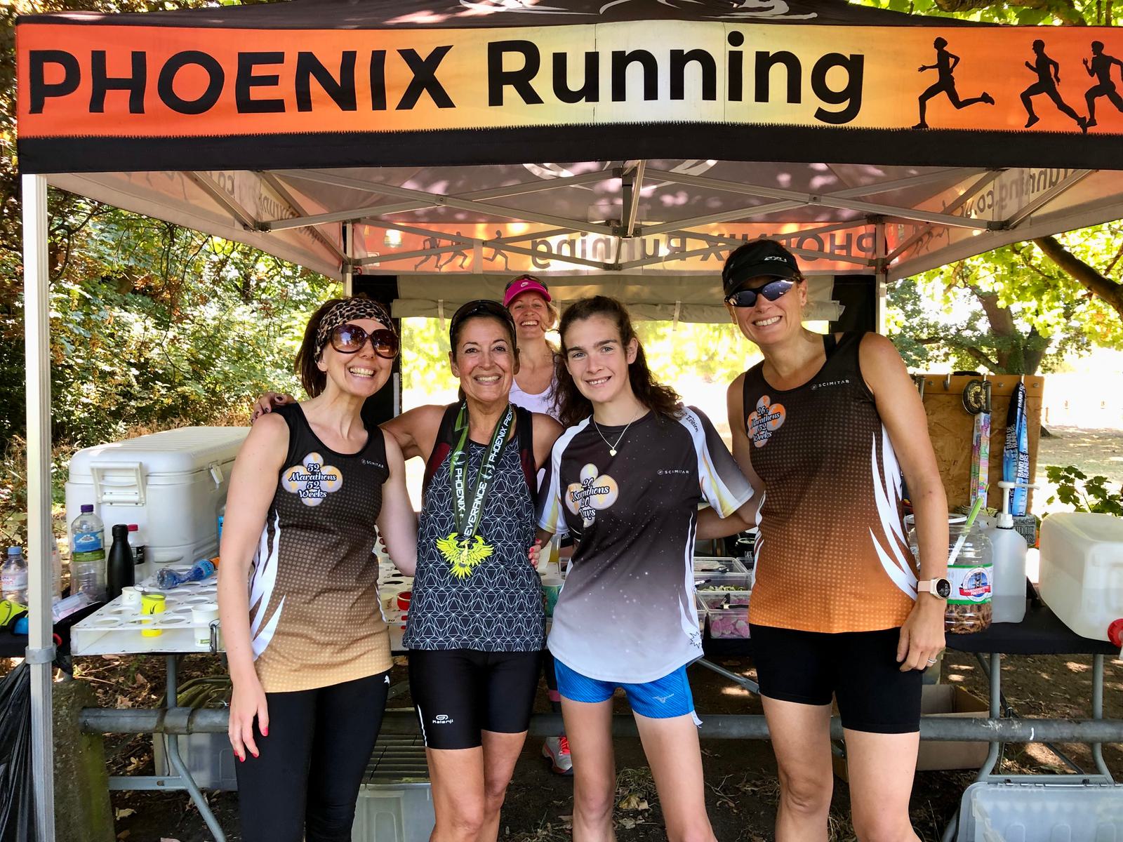 A group of five people wearing athletic gear stand smiling under a "PHOENIX Running" tent. One person wears a medal, suggesting a race. The background shows leafy greenery and race supplies on a table.