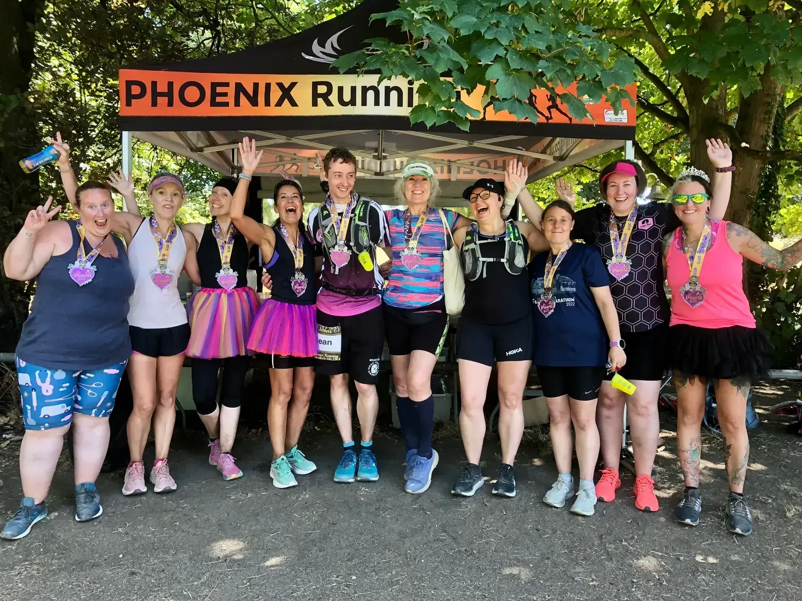 A group of nine people stand together posing and smiling under a tent that reads "Phoenix Running." They are wearing running attire and medals around their necks. Some have their arms raised in celebration. There are trees and greenery in the background.