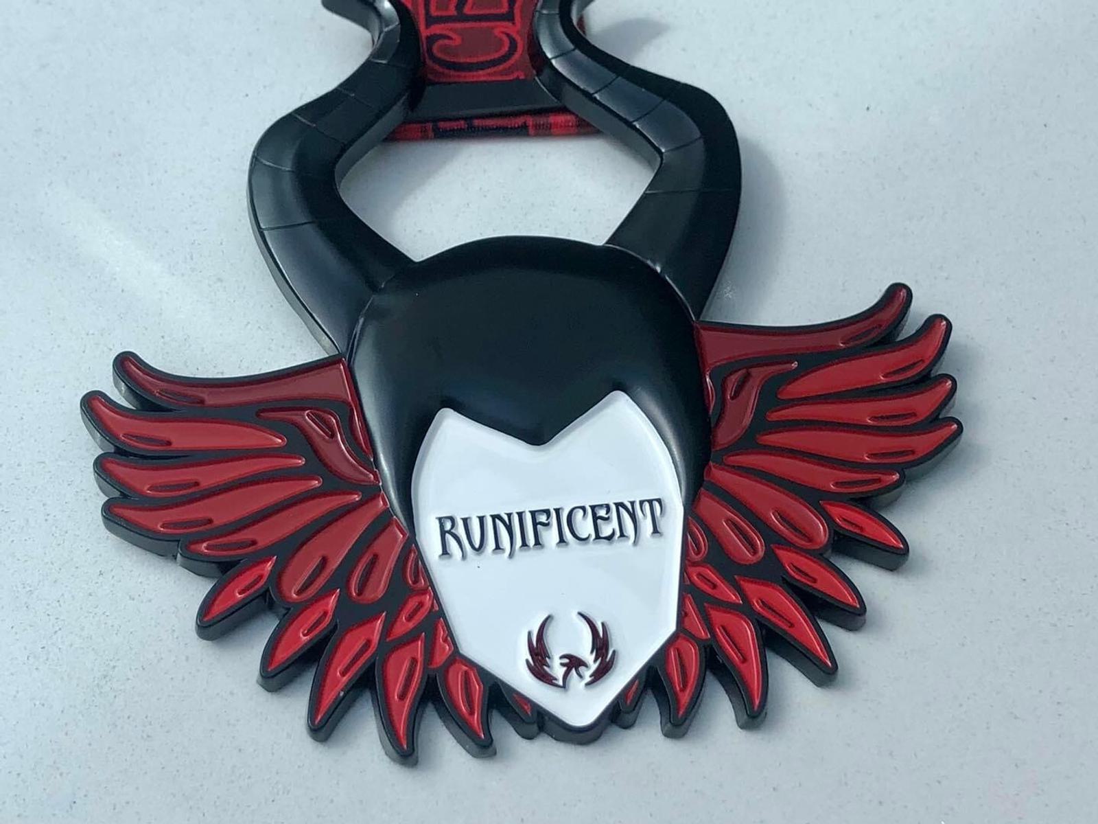 A black and red finisher's medal featuring large red wings, a stylized black horned headdress, and the word "RUNIFICENT" in capital letters. A small emblem at the bottom shows a stag head with antlers. The background is a smooth light gray surface.