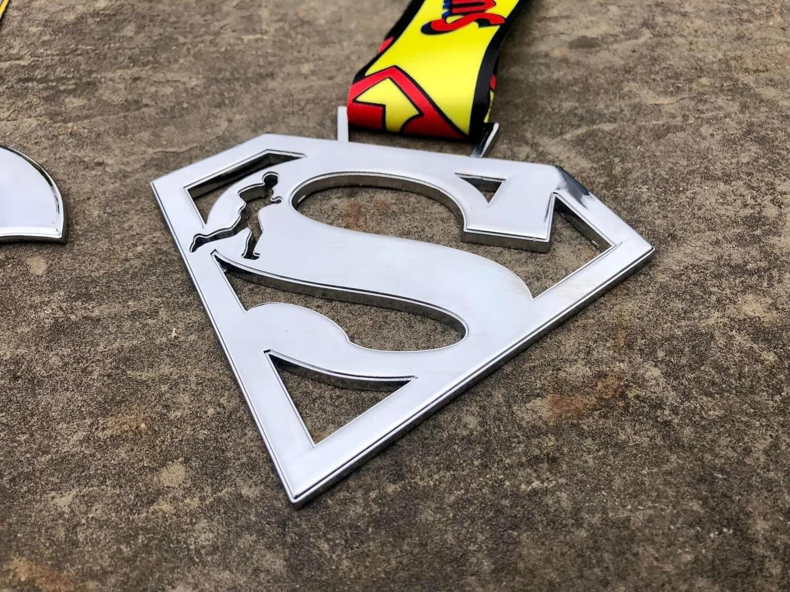 A medal with a large, metallic Superman "S" logo displayed on a textured stone surface. The logo is empty in the center, except for a small running human silhouette. The attached ribbon is colorful with red and yellow patterns.