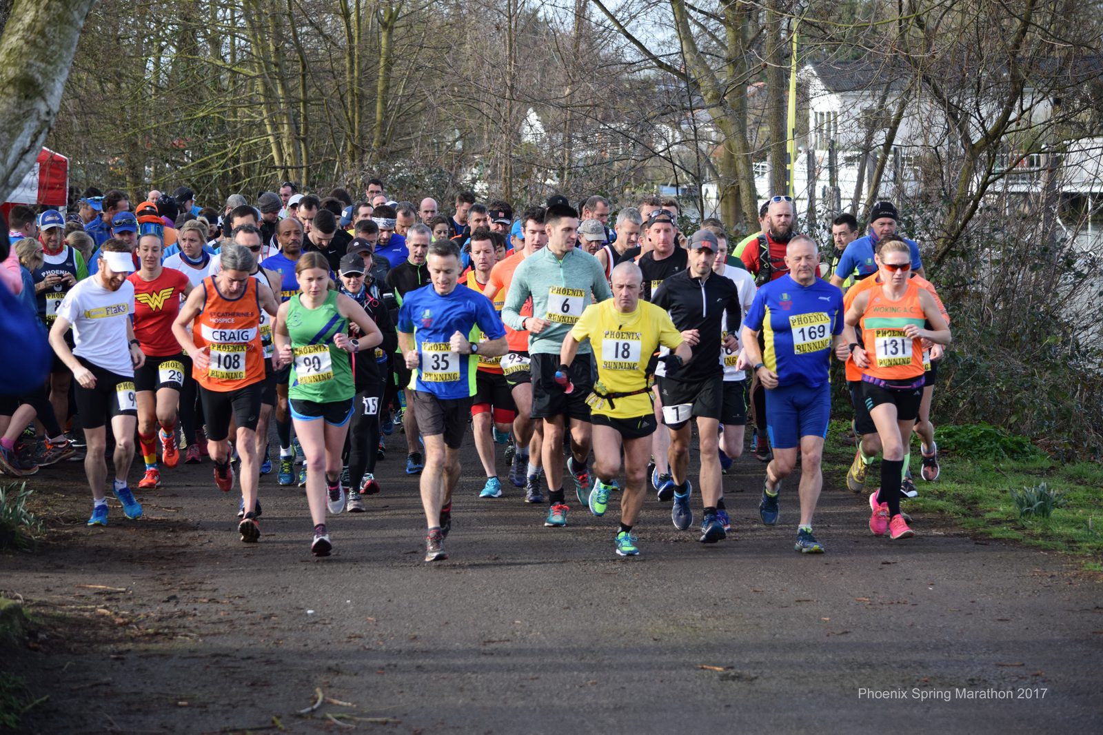 A diverse group of runners wearing numbered bibs and sports attire participate in an outdoor marathon, running on a path surrounded by trees and greenery. Several runners are leading the pack, while others follow closely behind. The atmosphere appears energetic and competitive.