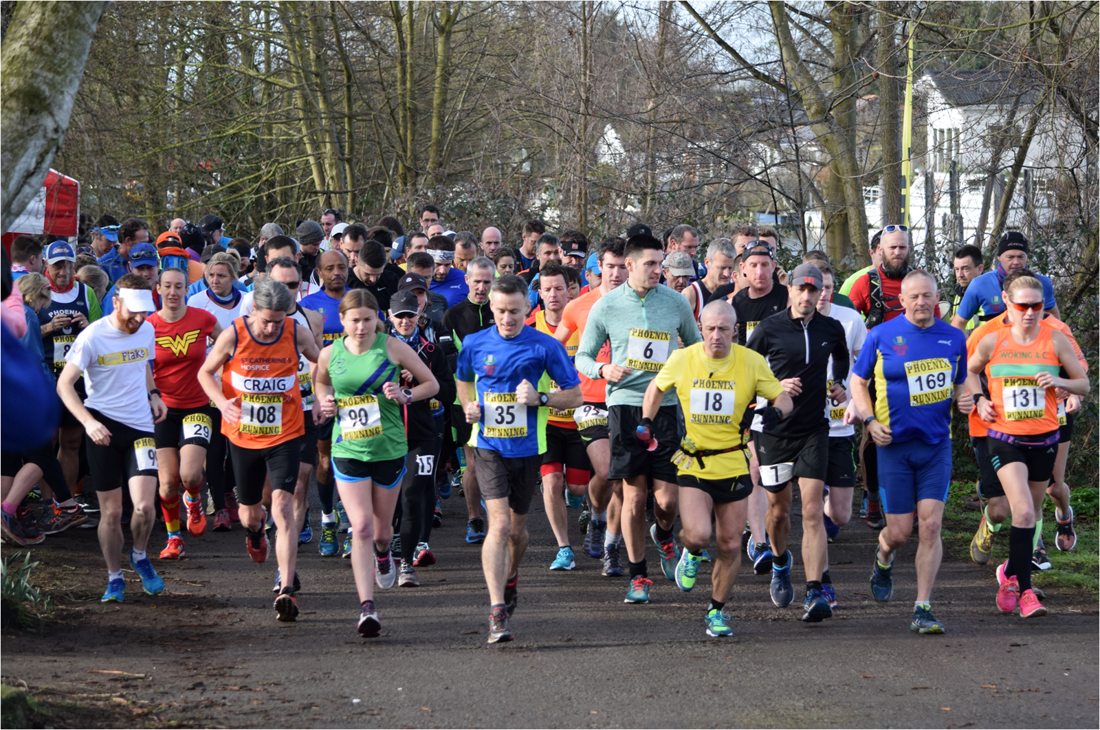 A large group of runners participating in a race, wearing various colorful athletic outfits and bib numbers, are seen at the starting point on a path surrounded by trees. The atmosphere is energetic as they begin their run.