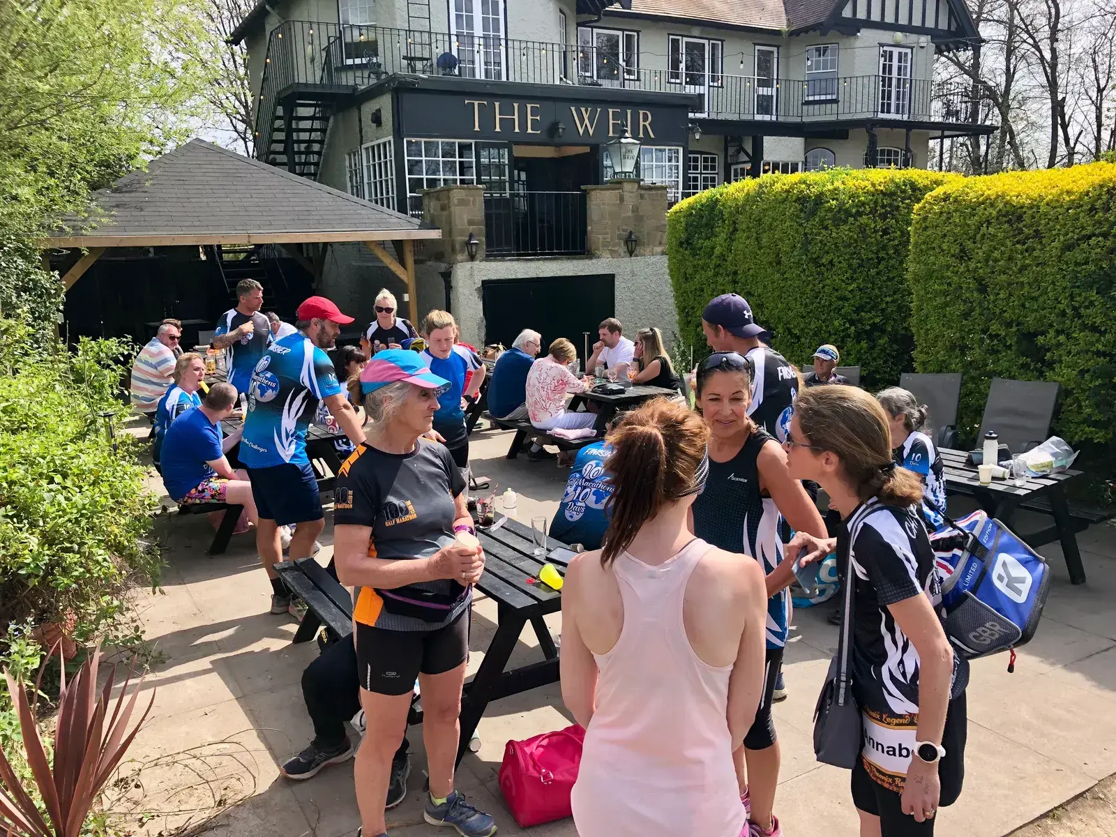 A group of people in activewear gather outdoors at picnic tables near "The Weir" pub. Some are standing and chatting while others are seated. The setting appears casual and sunny, with greenery and the pub building in the background.