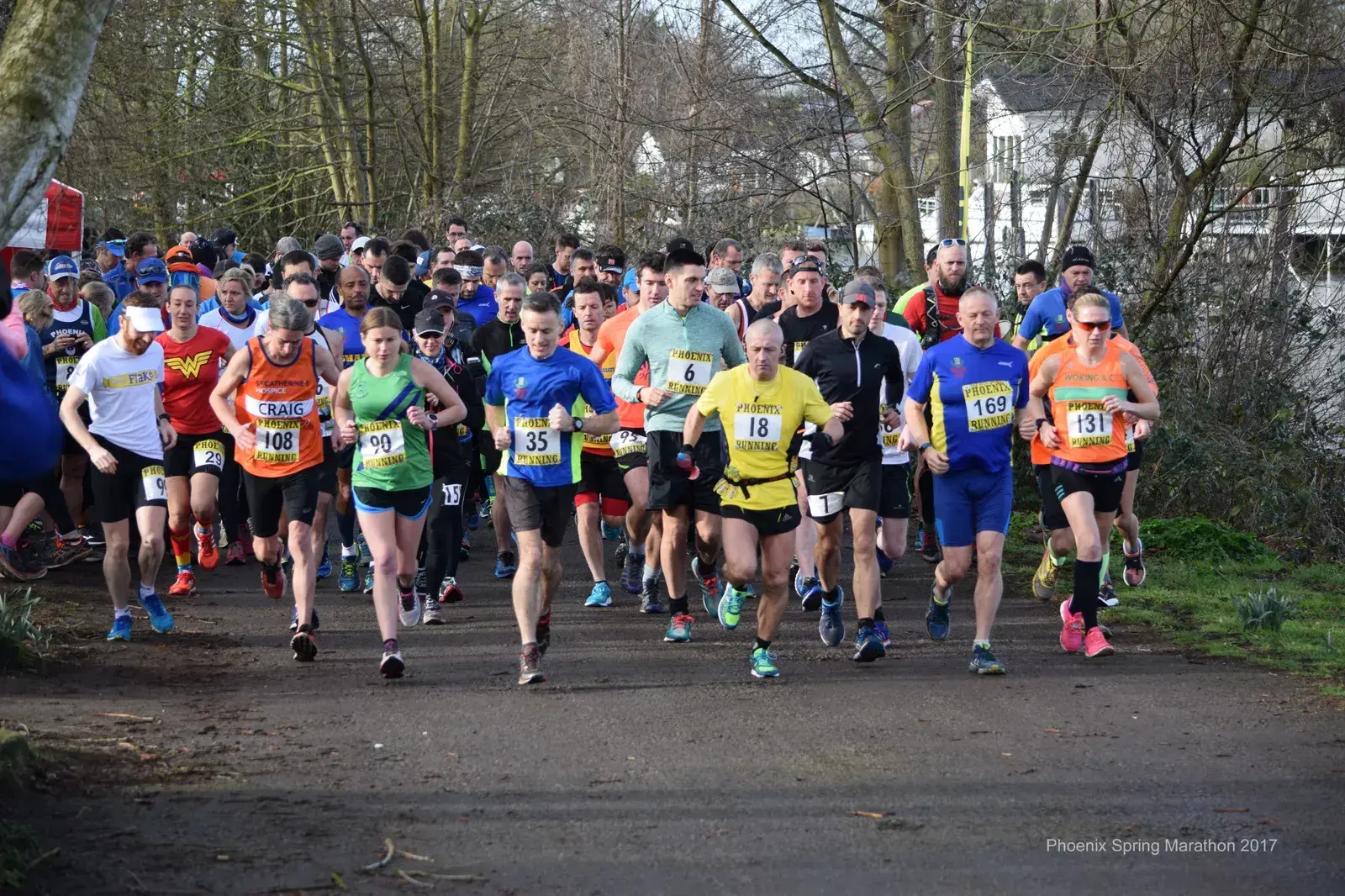 A large group of runners, each wearing numbered bibs, begin a marathon on a tree-lined path. Some runners are in brightly colored athletic wear, while others sport hats and sunglasses. Spectators can be seen in the background on both sides of the path.