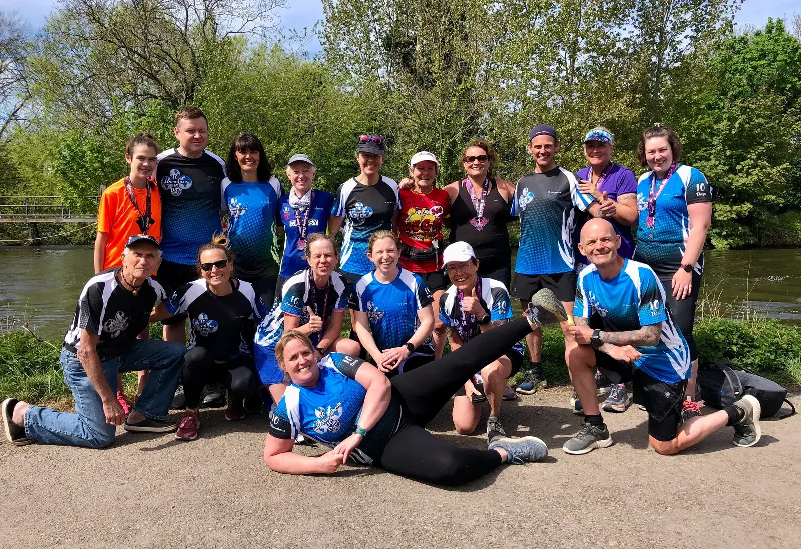 A group of sixteen people are posing outdoors after a run, dressed in athletic gear and wearing medals. They are standing in front of a body of water with trees and greenery in the background. Some are kneeling or seated in the front, smiling and looking happy.
