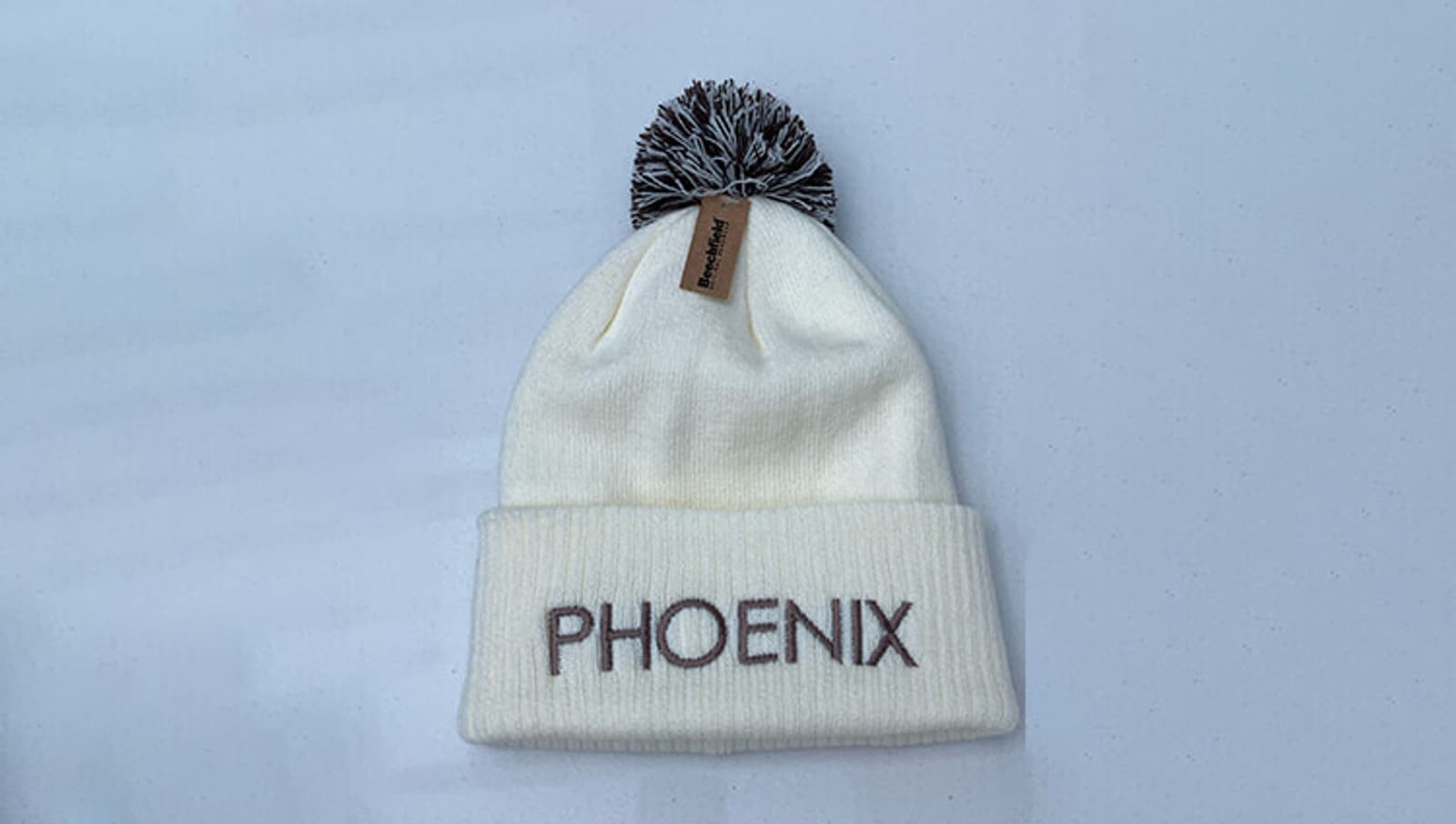 A white knit beanie with a gray and white pom-pom on top. The word "PHOENIX" is embroidered in large gray letters on the folded brim, and a small brown tag is attached near the pom-pom.
