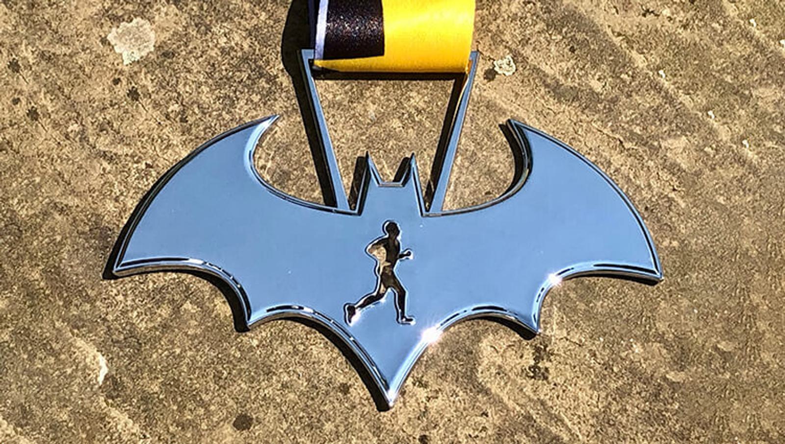 A metal medal shaped like the Batman logo with a small silhouette of a running person cut out in the center. The medal is attached to a yellow and black ribbon and is placed on a stone surface.