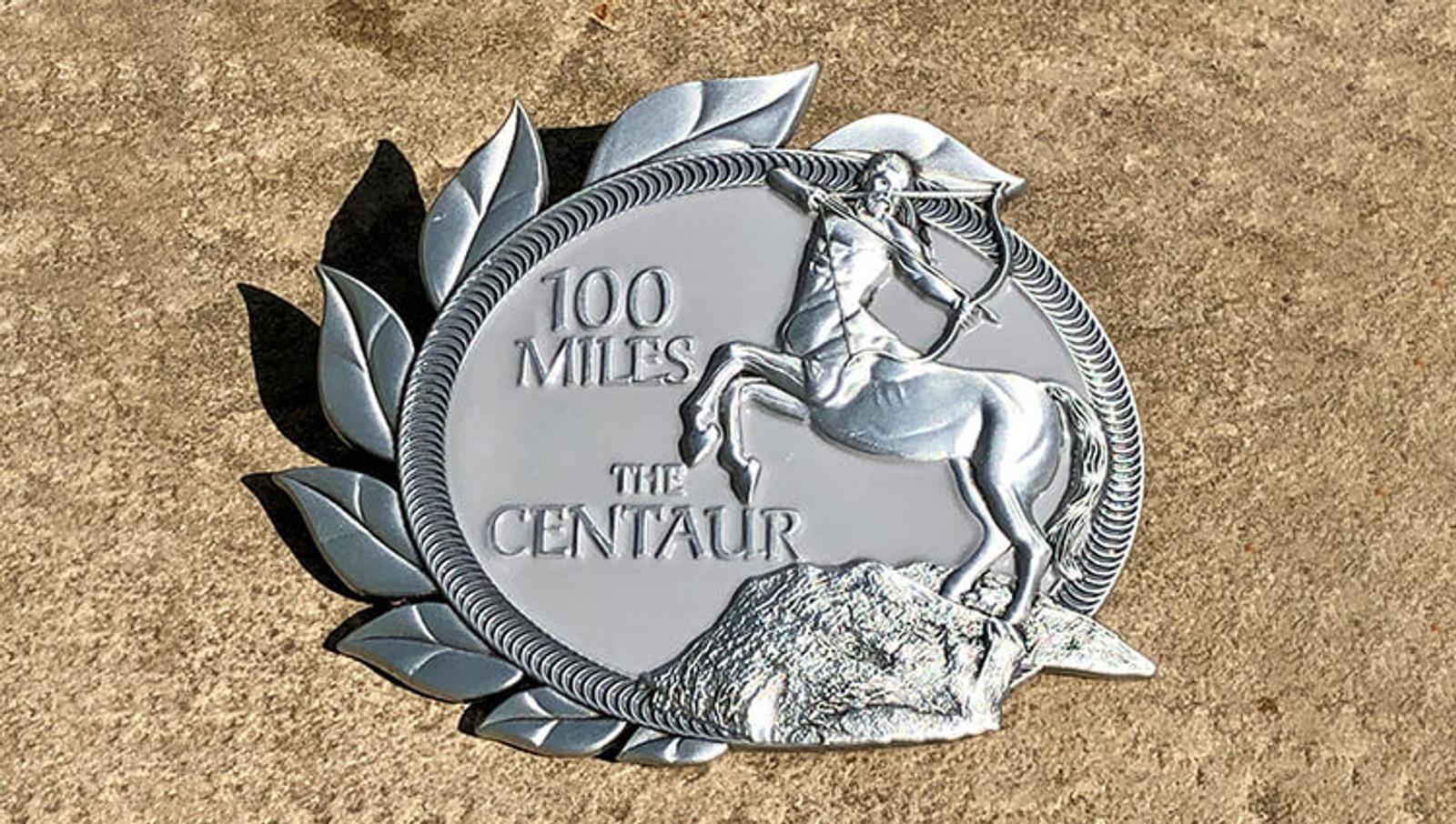 A silver medal with a relief of a centaur archer, surrounded by a laurel wreath. The text reads "100 MILES THE CENTAUR." The medal is placed on a concrete surface.