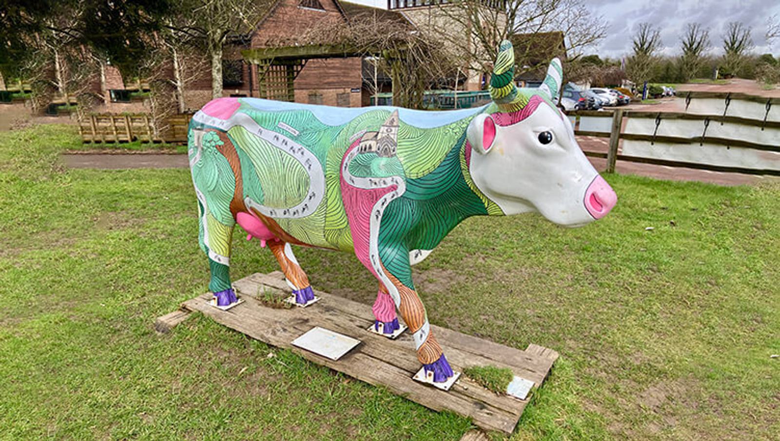 A colorful cow sculpture stands outdoors on grass. It features vibrant patterns with various colors, including green, pink, and purple. Its face is white with pink accents on the ears, eyes, and nose. Barn-like buildings are visible in the background.