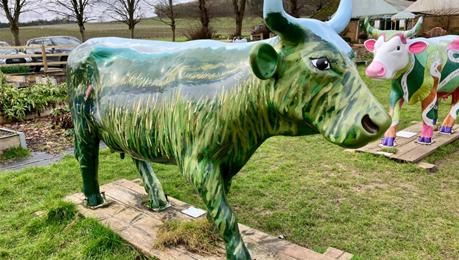 A life-sized cow sculpture painted with green grass-like patterns stands on a wooden platform outdoors, with another colorful cow sculpture and rural scenery in the background.