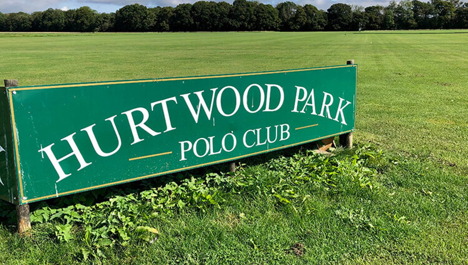 A large green sign stands on a grassy field with the text "Hurtwood Park Polo Club" written in white letters. The background features a row of trees under a clear sky.